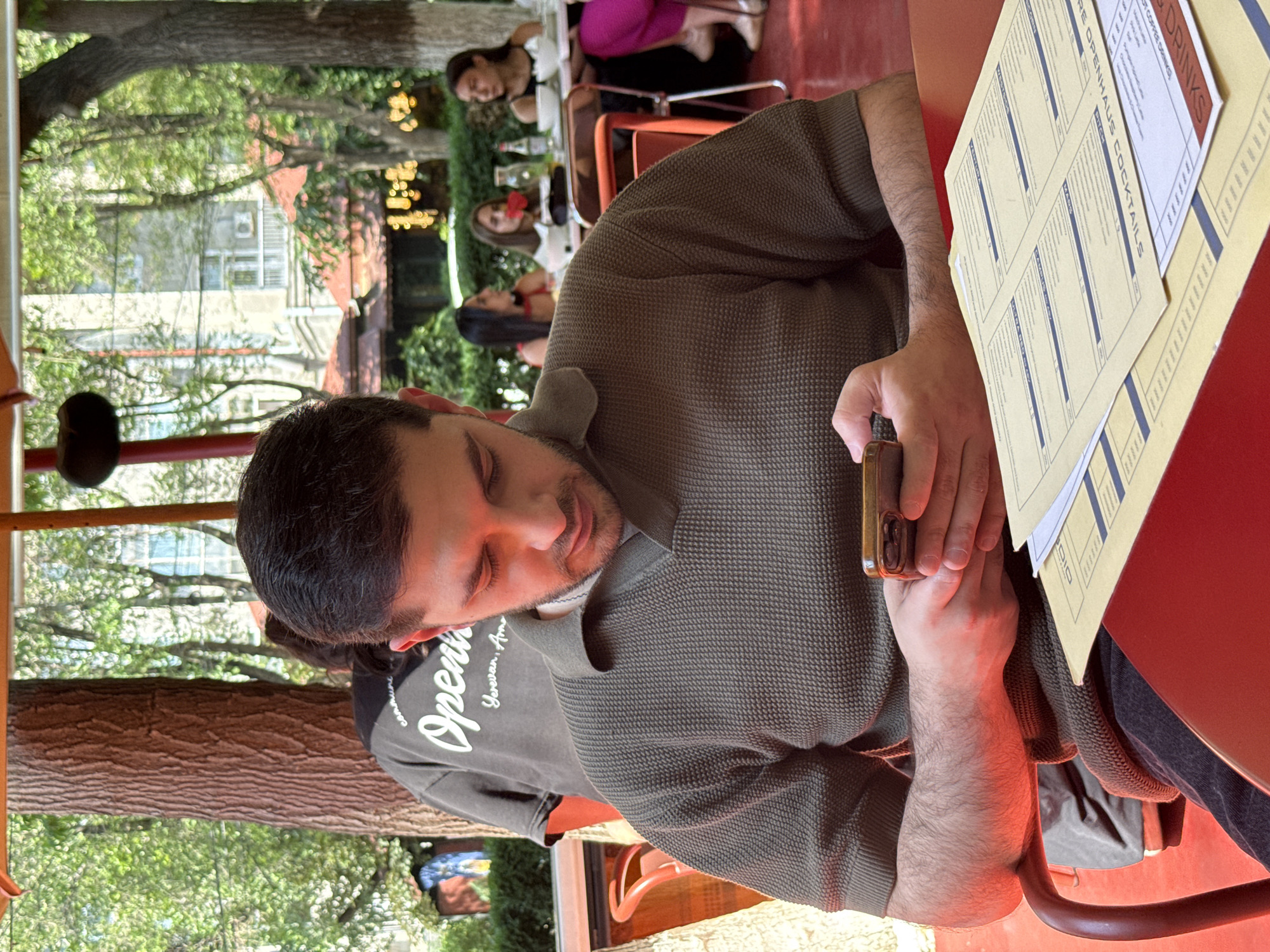 In Yerevan, Armenia, a man is seated at a red table in an outdoor seating area of a restaurant, engrossed in his smartphone. He is wearing a dark olive-green textured polo shirt. Several menus are spread out on the table in front of him. In the background, other patrons are visible at tables, and lush green trees and buildings are present. A tree trunk is prominently positioned to the left of the man.