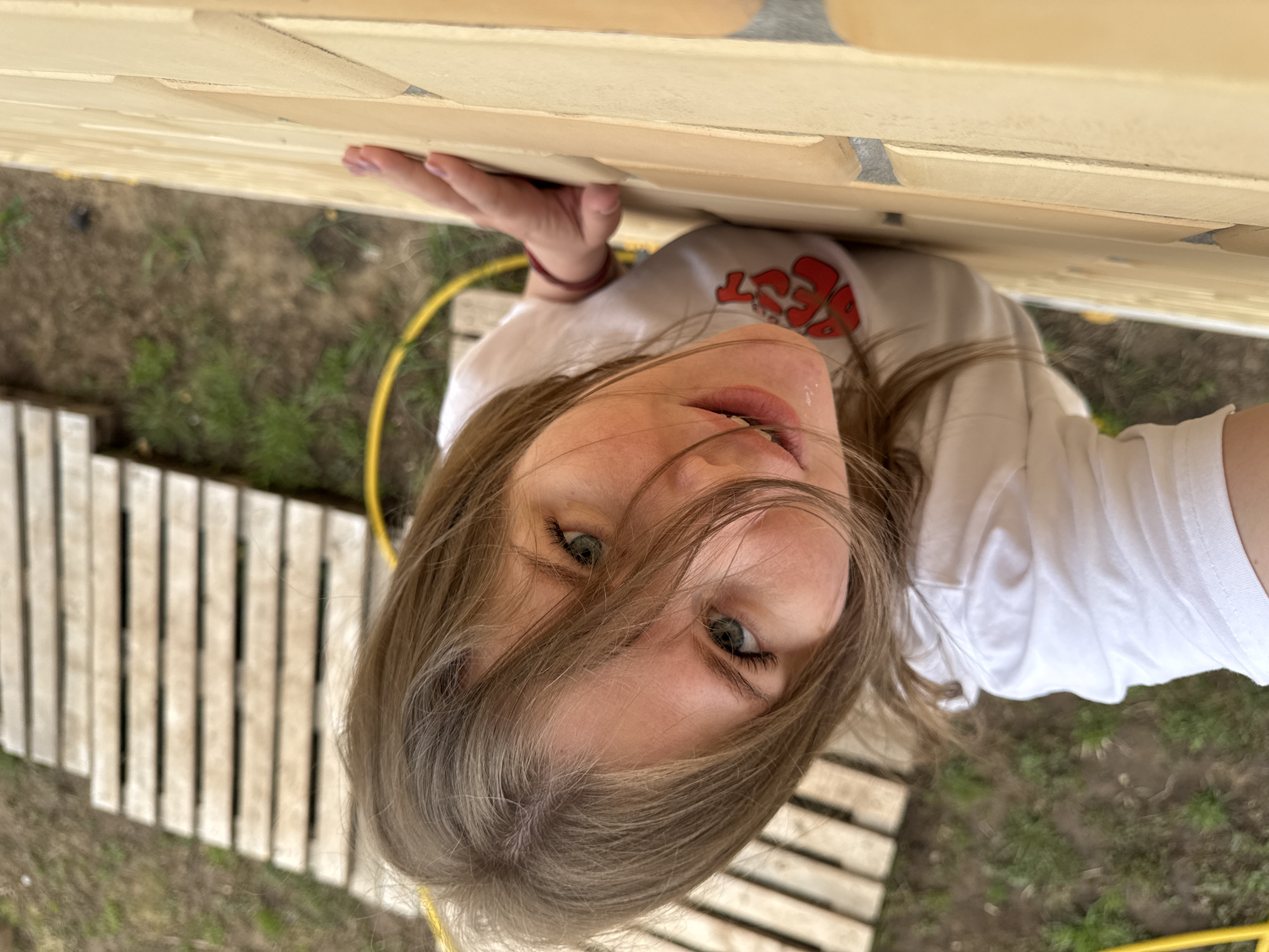 A young woman is captured in a high-angle shot, looking directly up at the camera. She has long, light brown hair, slightly messy and falling around her face, and blue-green eyes. Her lips are slightly parted. She is wearing a simple white short-sleeved t-shirt with a red and white graphic design on the chest; visible letters appear to form "BEST" in a stylized font. Her right hand, with light nail polish, is seen holding onto the edge of a light-colored wooden structure or wall, which appears to be directly above her.
The setting is outdoors, possibly in a backyard or garden in Gornyi (Dvurechenskoe S/P), Russia. The ground below her is a mix of bare earth and patches of sparse green grass. A bright yellow garden hose snakes across the ground in the mid-ground. To her left and right, blurred out, are sections of light-colored wooden pallets or slatted structures lying flat on the ground. The lighting suggests it is daytime, with soft, even illumination. The overall impression is a casual, relaxed moment, possibly a selfie taken from a unique perspective while lying down or looking up from a low vantage point.