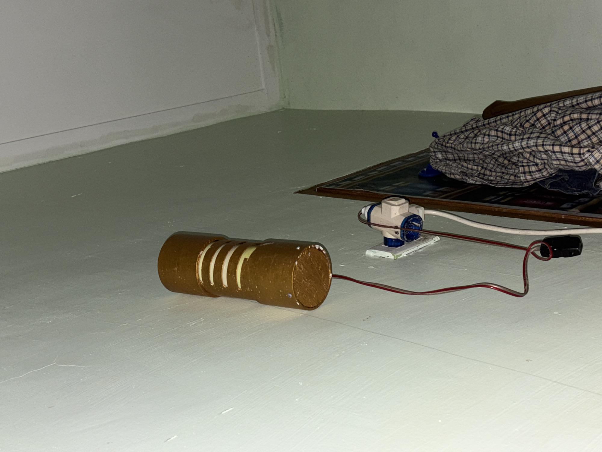 A homemade, cylindrical lamp with horizontal stripes of light and dark gold is mounted on a light green wall. A red wire extends from the bottom of the lamp to a small black object, possibly a transformer. Below the lamp, a white electrical socket is mounted on the wall, with a blue and white connector plugged into it. Two wires run from the socket, one red and one white. To the right of the electrical socket, a framed object, possibly a mirror or a picture, leans against the wall. A plaid shirt is draped over the top of the framed object. The ceiling is visible in the upper right corner of the image and appears to have water stains. The image is taken from a low angle, looking upwards towards the lamp and ceiling. The location is identified as Tupah, Indonesia.