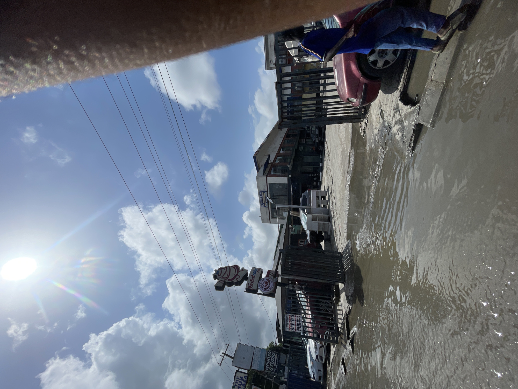The image shows a flooded street in Ado, Nigeria. The water level is high enough to cover the sidewalk and partially submerge the road. A red car is partially in the water. A motorcycle rider wearing blue pants and a blue shirt is on a vehicle next to the car. Several buildings are visible in the background, including one with a "Taster's" sign. There is a sign in the front of the building advertising a "HANDYMAN NEEDED." Power lines run across the sky, which is partly cloudy with the sun shining through. A portion of a light-skinned human arm is in the top left corner of the picture.
