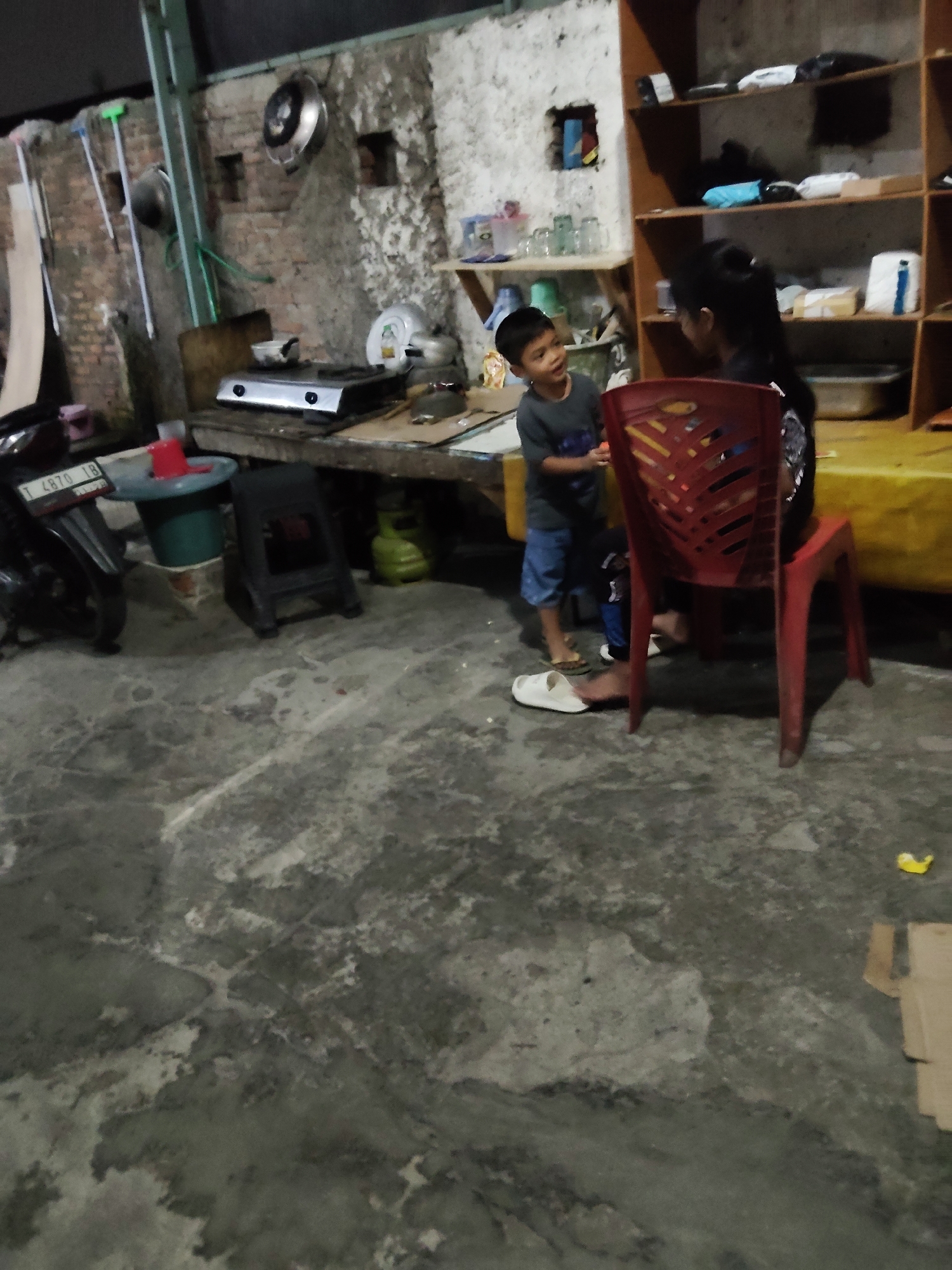 The image depicts an outdoor, covered area, likely a kitchen or storage space, in Mekarmukti, Indonesia. A young boy, wearing a dark t-shirt and blue shorts, stands on the concrete floor, facing and interacting with an adult woman. The woman, with her dark hair pulled back, is seated on a red plastic chair. She is wearing dark clothing. The environment is cluttered with various objects. To the left, a motorcycle is parked, and a green stool is visible. A gas stove is on a wooden counter. Shelving units are built into the wall, containing items like containers and what appear to be cleaning supplies. The walls are rough and unfinished brick, with some areas showing plaster. The floor is concrete with visible cracks and stains. The lighting appears to be artificial, suggesting it might be evening or a dimly lit indoor space.