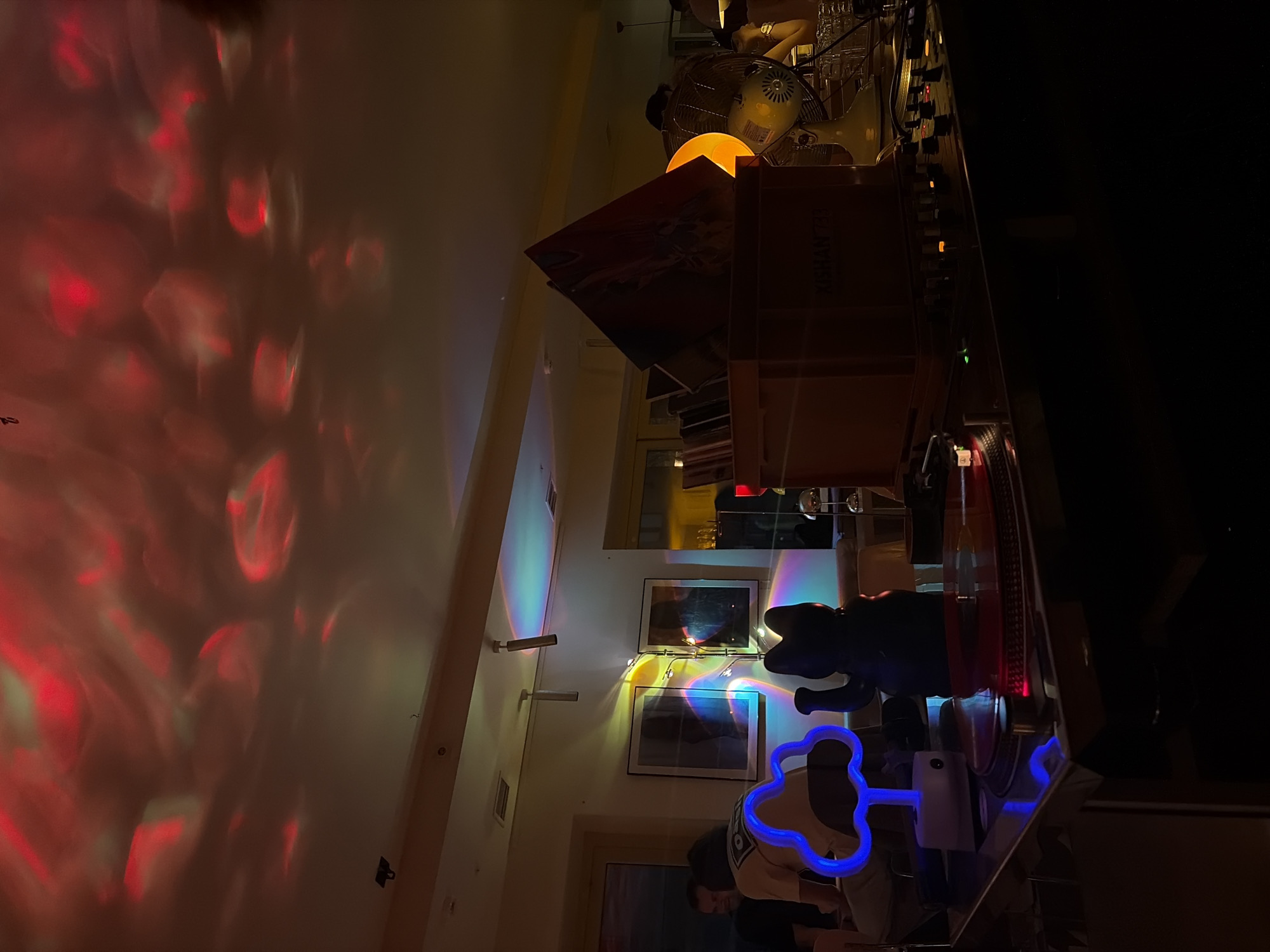 The image shows an interior scene. Red light patterns are projected on the left wall. Along the far wall are two framed pictures hung one above the other, illuminated by string lights. A blue neon light in the shape of an abstract object is visible, along with a figure of an elephant. Stacks of records and a mixing console are positioned on a surface. A person is visible in the lower center of the frame.