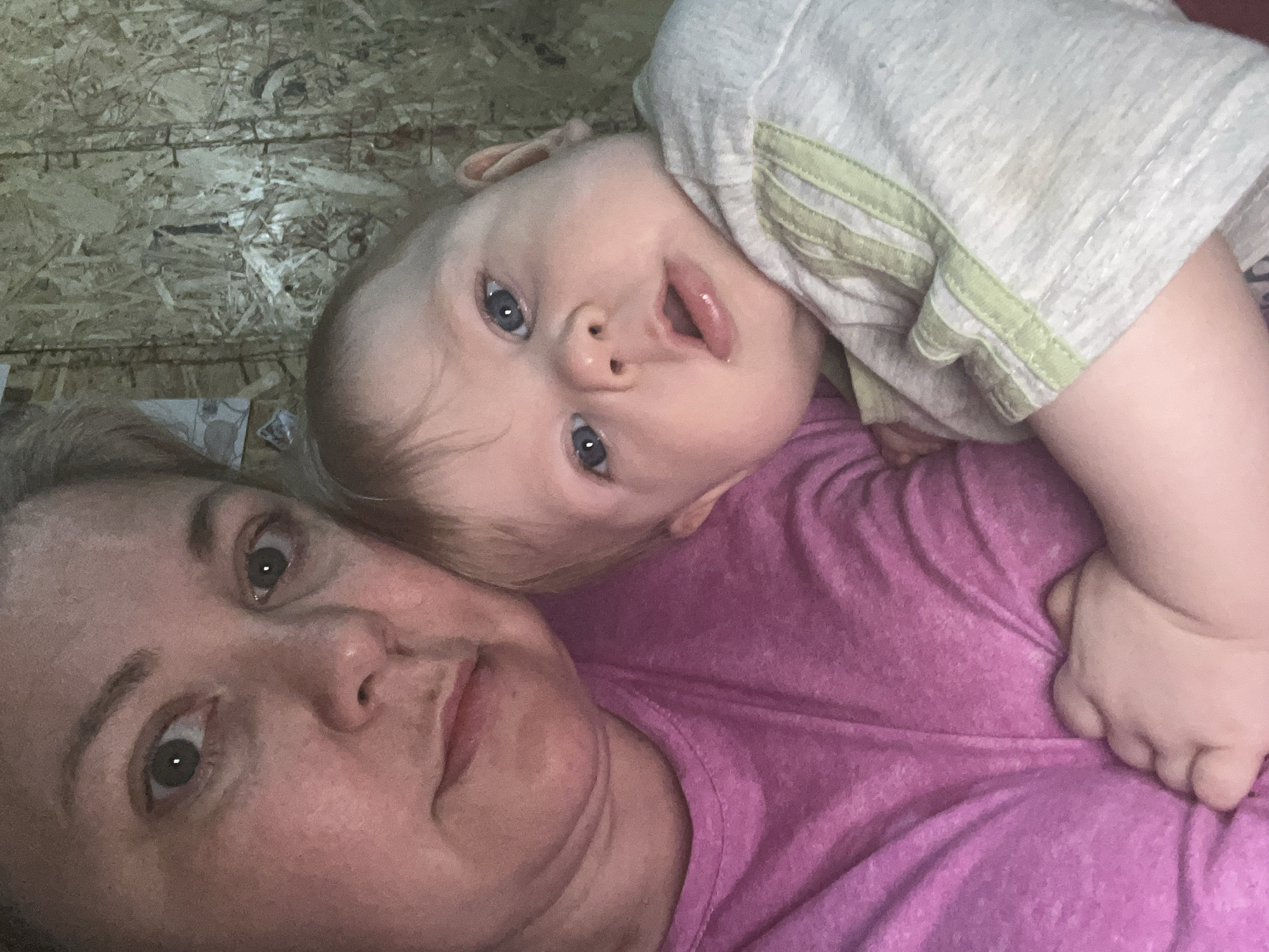 A selfie image shows a woman lying down with a baby resting on her chest. The woman is wearing a pink shirt and looks directly at the camera. The baby, wearing a light-colored shirt, looks up with its mouth slightly open. The background is a wooden surface. No specific location or city is discernible from the image.