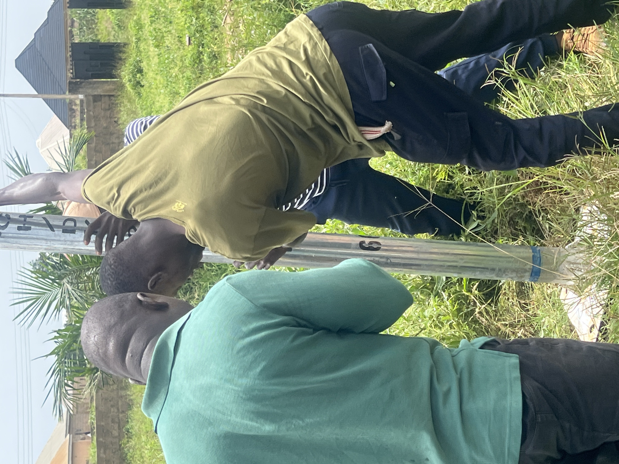 Three individuals are partially visible, engaged in an activity around a metal pole in an outdoor, grassy environment. One person, wearing a teal polo shirt, faces away from the camera, their back to the viewer. To their right, another person in a dark olive green t-shirt is leaning over the pole, their arms extended as if working on it. Their dark pants have cargo pockets. A third individual, mostly obscured, is wearing a striped shirt and dark pants. The scene appears to be in a moderately vegetated area, possibly a field or undeveloped lot, with some structures and palm trees visible in the background. The lighting suggests it is daytime. The metal pole has markings on it, including what appears to be the number "6". The overall impression is one of manual labor or installation work. The image does not provide specific visual cues that would allow for definitive identification of the location as Jos, Nigeria.