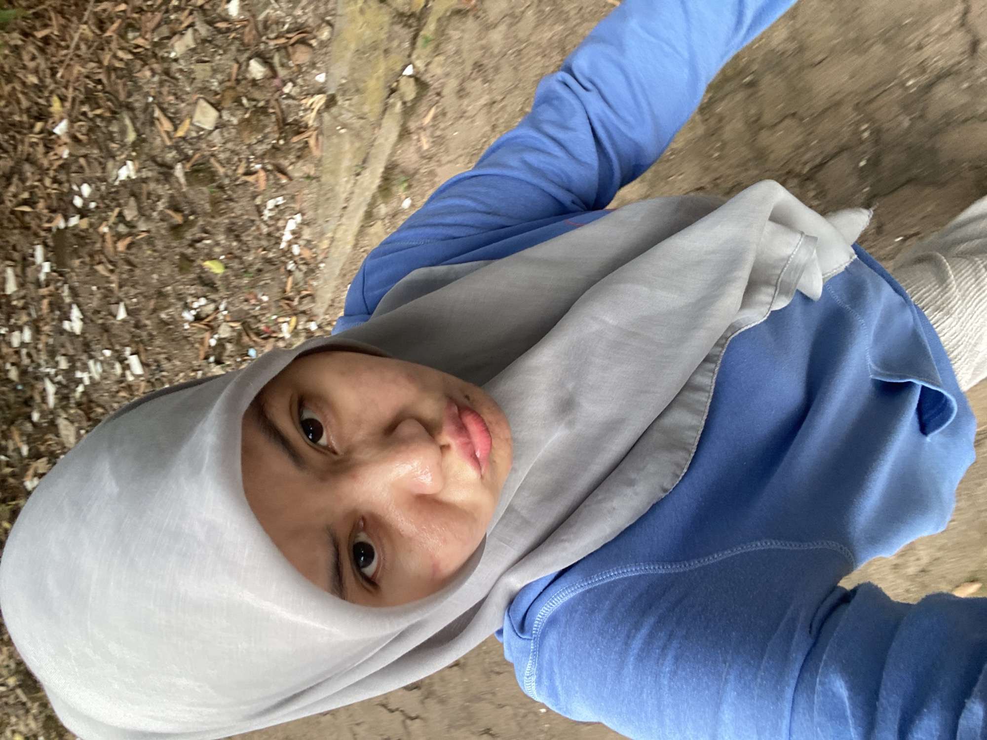 A woman wearing a light gray hijab and a blue long-sleeved shirt is captured in a selfie. Her face is close to the camera, with her lips pursed. The background appears to be an outdoor area with dirt, fallen leaves, and small scattered white objects, suggesting a natural or unkempt environment. The specific location in Banjarbaru, Indonesia, is not discernible from the visual elements alone.