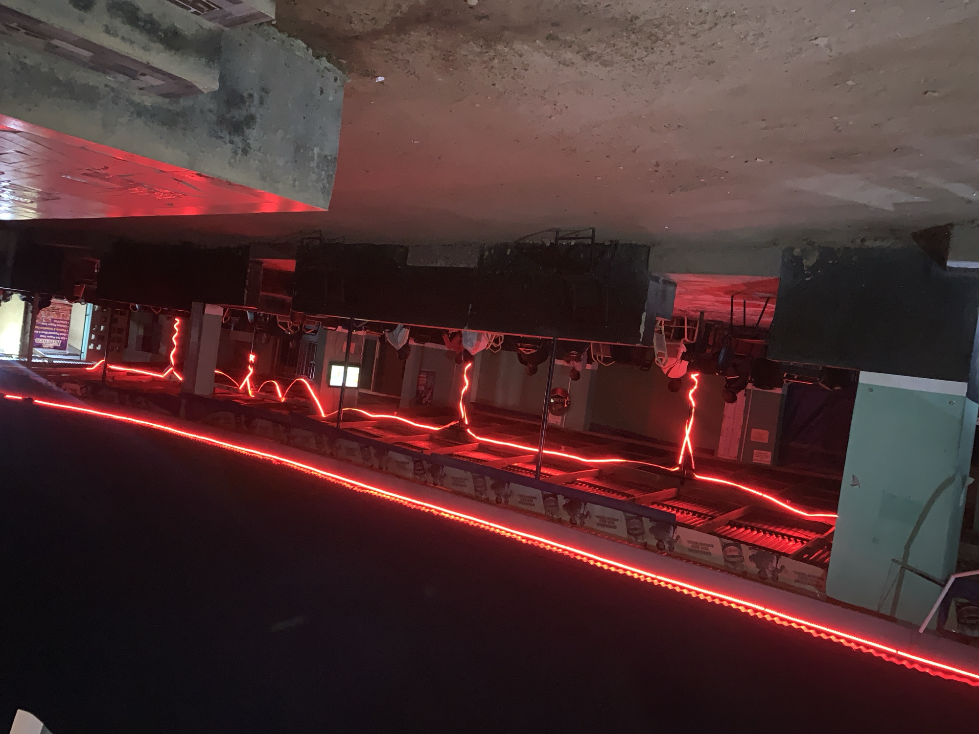 The image depicts an upside-down view of what appears to be a platform at the New Stadium Train Station in New Stadium, Nigeria. Red neon lights are prominent, outlining the platform edges and illuminating architectural features. The platform appears to be illuminated overhead and underneath with red lights. There are no people visible in the image. The environment appears to be an exterior train platform with cement and concrete structural elements.