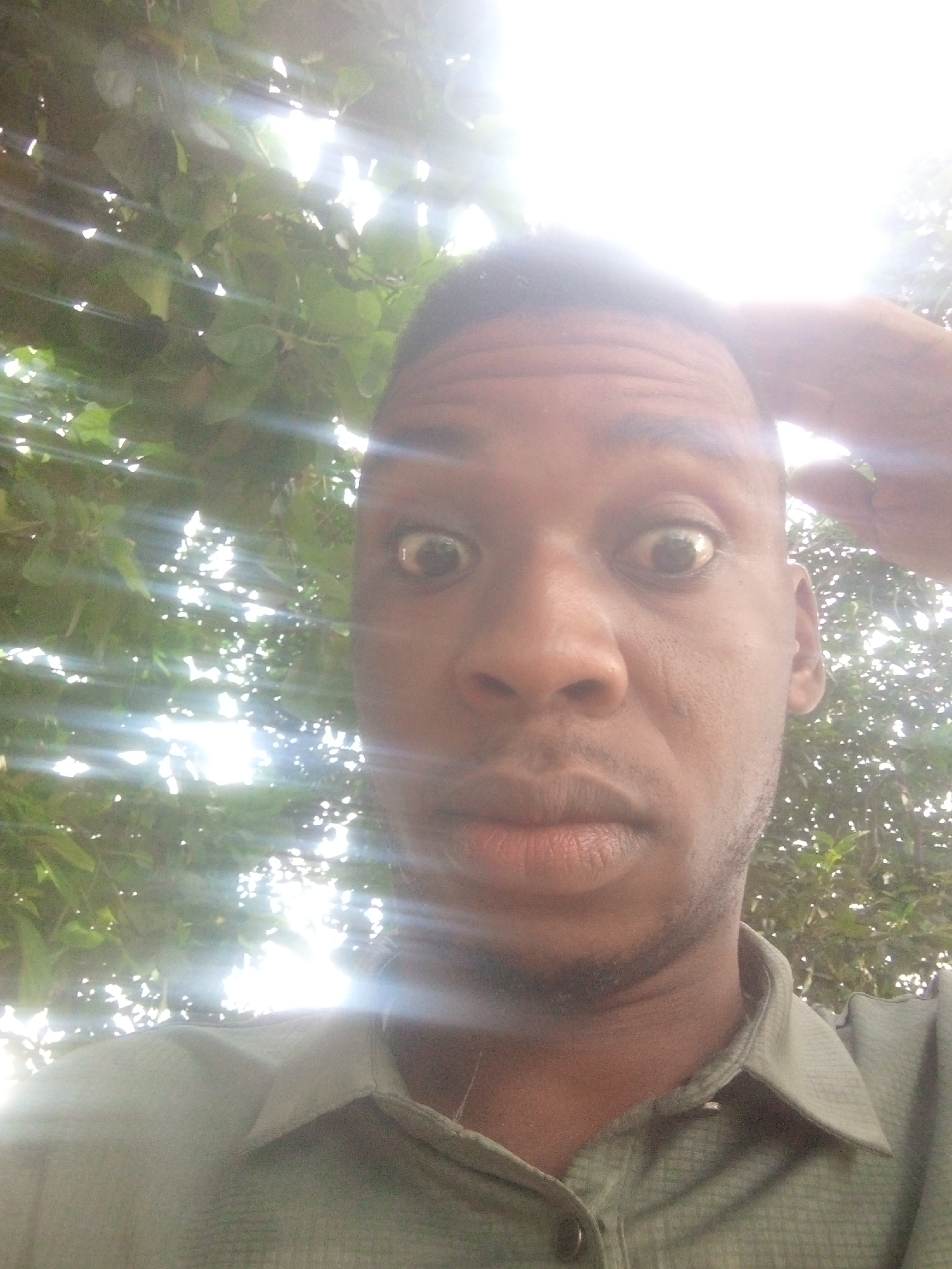 A close-up selfie of a Black male with wide eyes is taken in an outdoor, sunlit environment. The subject is wearing a collared shirt. The background includes trees and bright sunlight creating strong lens flares. The location is unspecified as 'Unknown, Unknown'.
