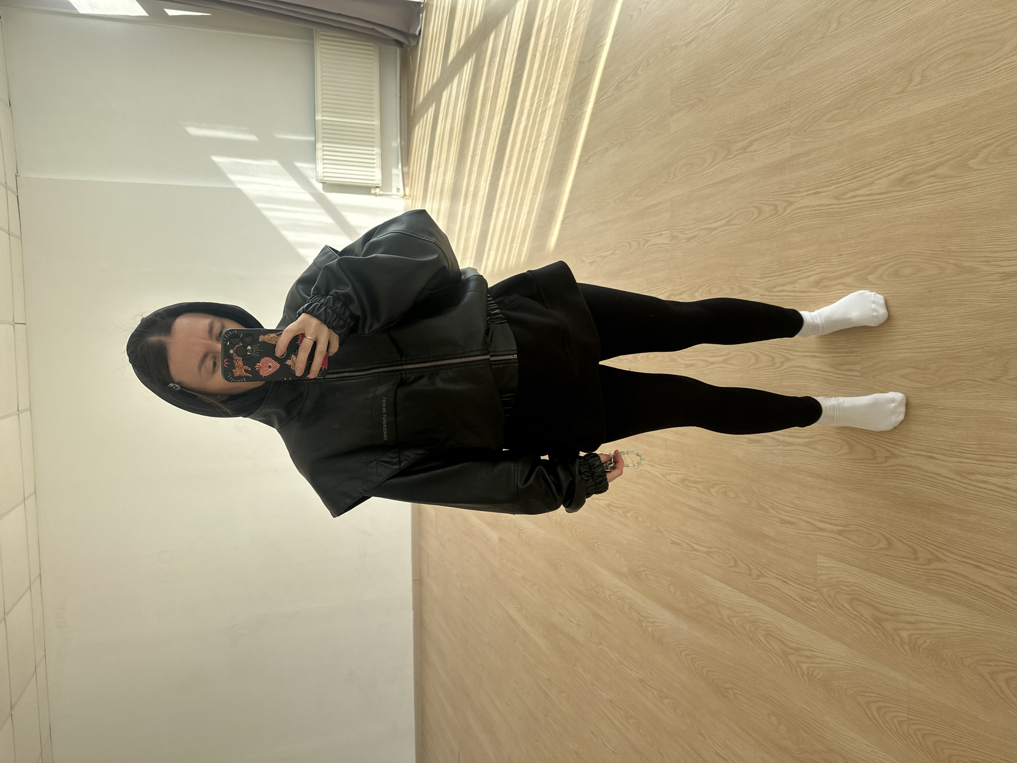 A person is taking a selfie in a room with light-colored wooden floors and white walls. The person is wearing a black hooded jacket, black skirt or shorts, black leggings, and white socks. Sunlight streams through blinds in the background. The phone case features floral and animal motifs. The location is indeterminate without further information, but the request includes a city; therefore, the location is somewhere in Votkinsk, Russia.