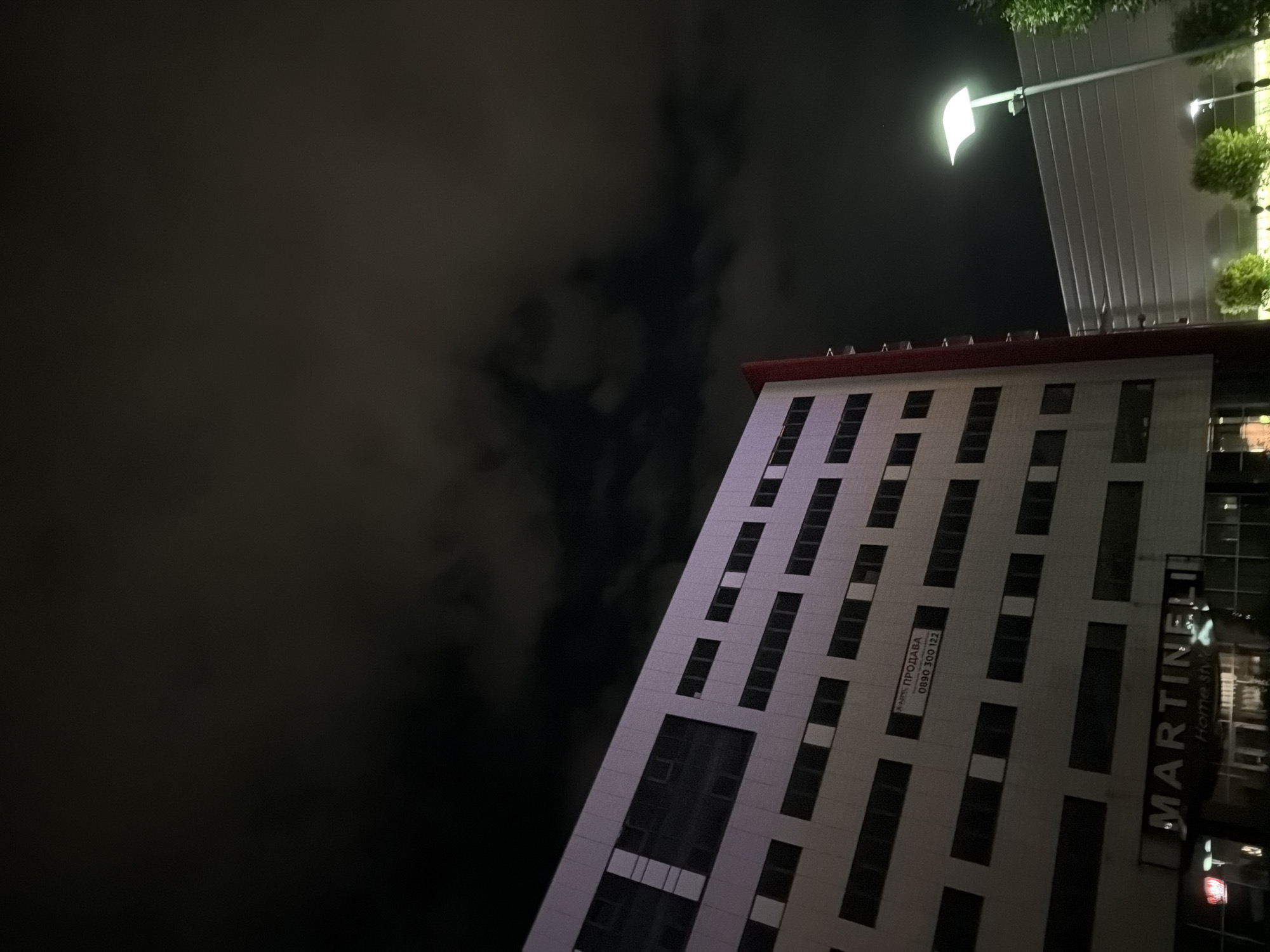 The image shows a modern building at night in Ruse, Bulgaria. The building is tall and appears to be made of light-colored panels with numerous dark windows. A red vertical element runs along the side of the building. A sign with Cyrillic text and a phone number is visible on the building's facade. At the bottom of the building, a lit sign reads "MARTINELLI Home style". In the background, another modern building is visible, illuminated by a street lamp. The sky is dark with some cloud cover, and there are some trees and greenery in the foreground. The overall scene suggests an urban commercial or residential area at night.