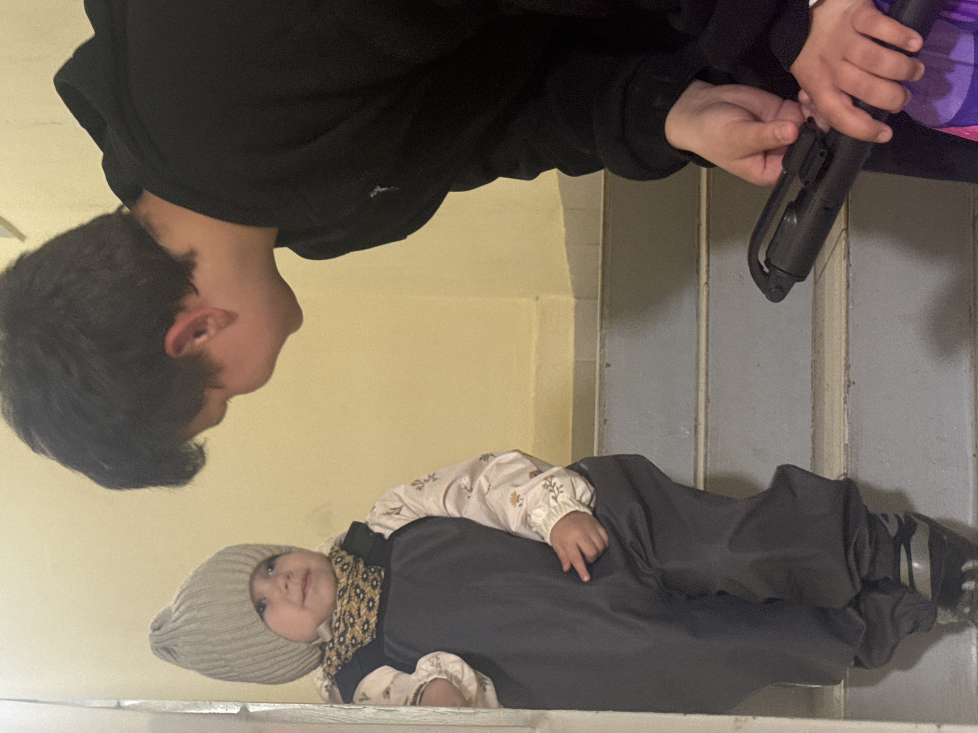 A child lies on their back wearing a grey knit hat, a floral print shirt, a patterned scarf, and dark grey overalls. A young person in a black hooded sweatshirt looks down at the child. A small pair of hands holds a black object near the edge of a staircase. The location is Chaikovsky, Russia.