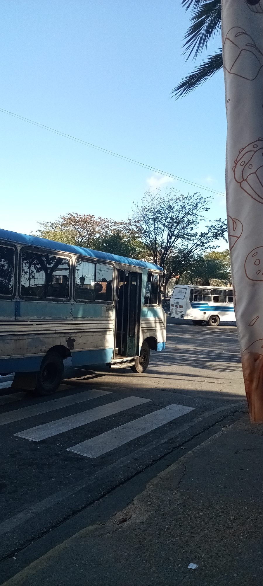The image depicts a street scene with two buses, one foregrounded and the other in the background, in the city of Unknown, Unknown. The foregrounded bus, painted in shades of blue and white, appears to be at a standstill on a zebra crossing. Its door is open, hinting at possible passenger disembarkation or boarding. The bus exhibits signs of wear and tear.
The background reveals another bus of similar color scheme, farther down the street. The environment includes trees in varying shades of green and a clear, bright blue sky. A partially visible pole with patterned fabric is present on the right side of the image. The street itself is paved with asphalt. The image suggests a daytime setting with natural light.
