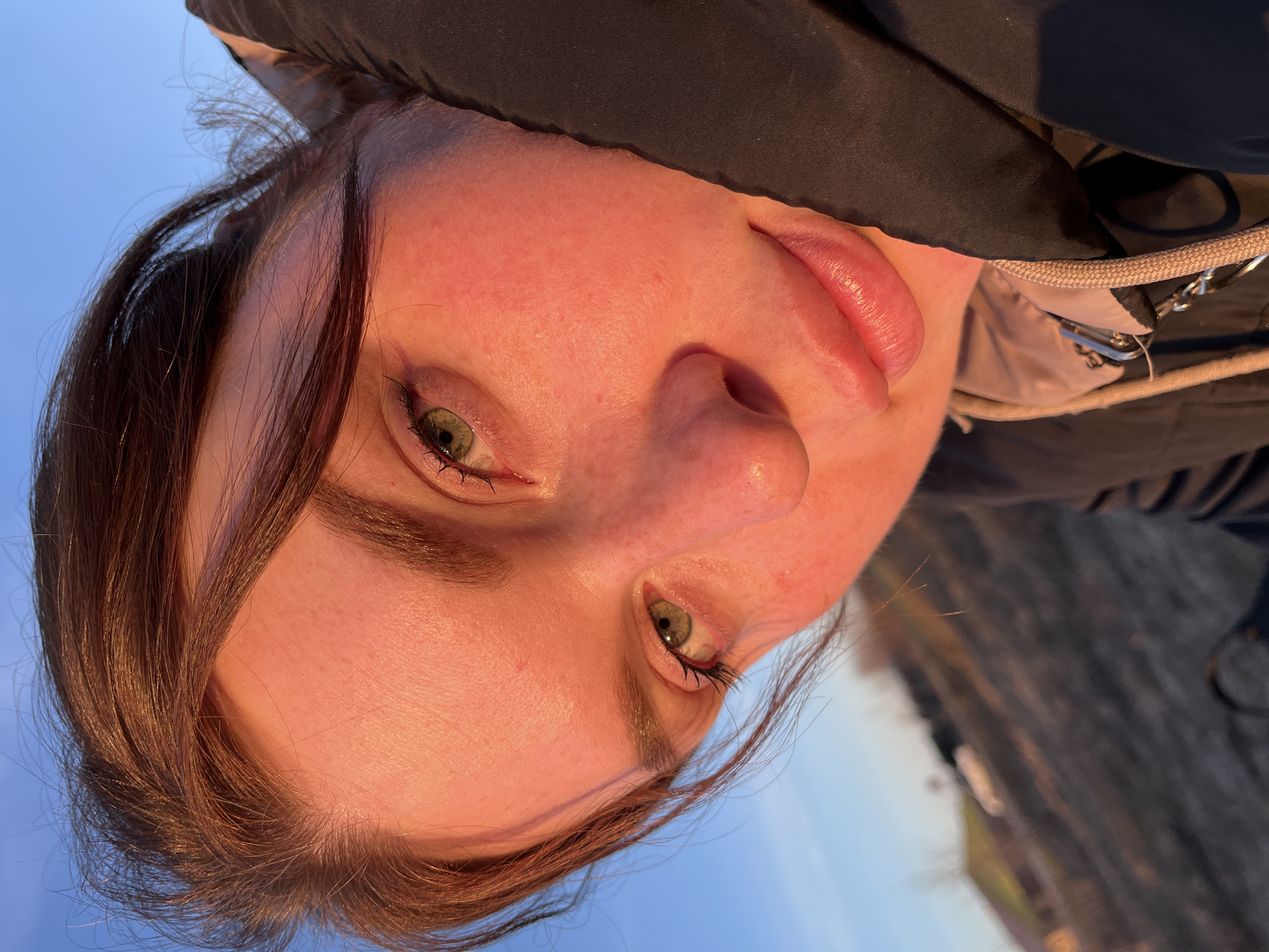 A close-up, rotated view shows a woman's face, with fair skin, brown hair, and green eyes. She is wearing a dark-colored jacket, partially visible around her head and shoulder. The background appears to be a blue sky near Павловка, Russia.