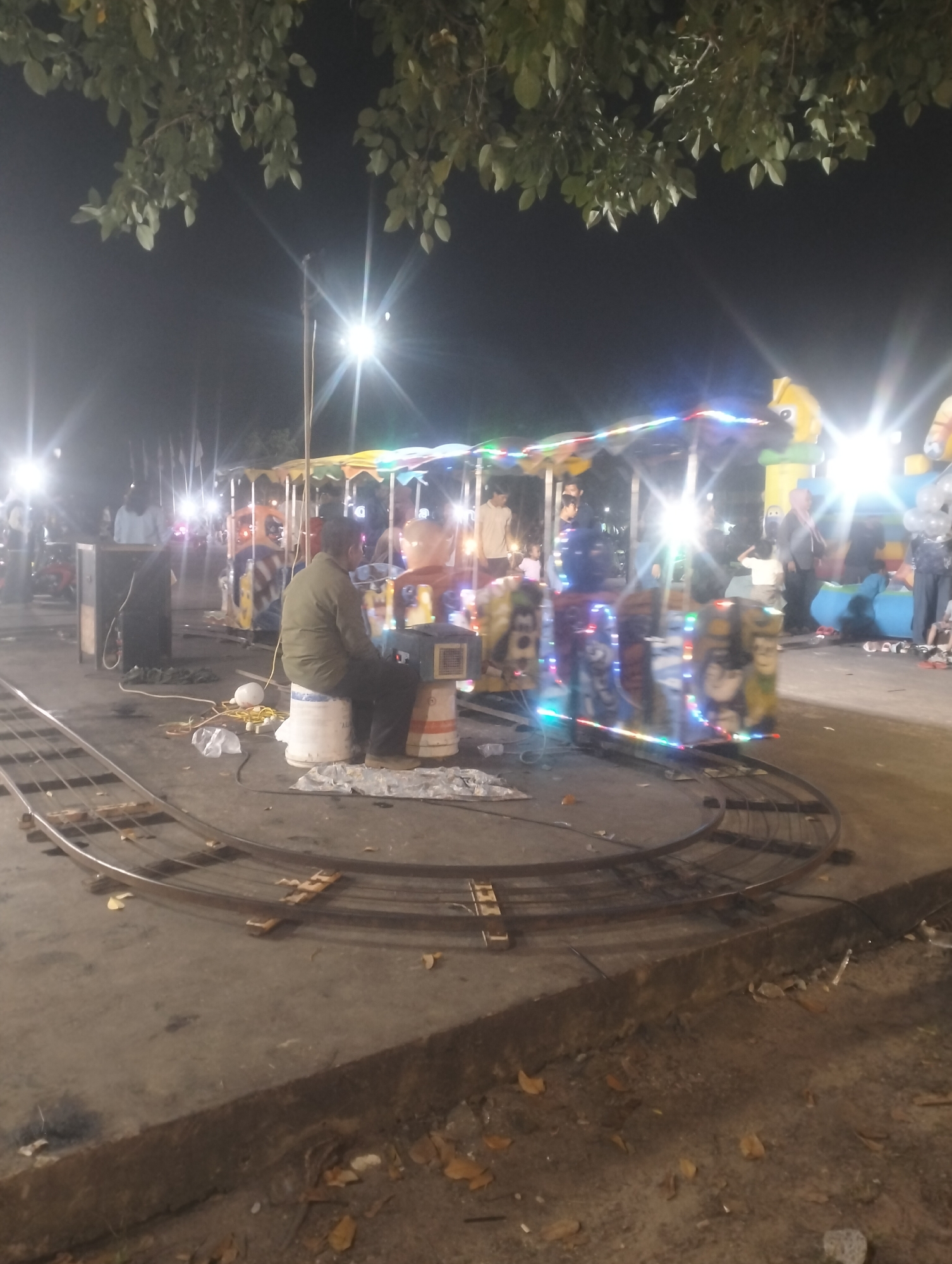 This is a night-time photograph of a small amusement park ride, likely a carousel or miniature train, situated outdoors on a paved surface, possibly in a public park or fairground in Dumai City, Indonesia. The ride is illuminated with colorful string lights, and in the background, there are more bright lights and the silhouettes of trees. A man in a green jacket is seated on a white bucket, facing away from the camera and towards the ride. Several other figures, including adults and possibly children, are visible in the background, suggesting the ride is operational or about to be. Metal tracks for the ride are laid out in the foreground, curving to the left. The ground is paved with concrete and scattered with fallen leaves, indicating an outdoor setting.