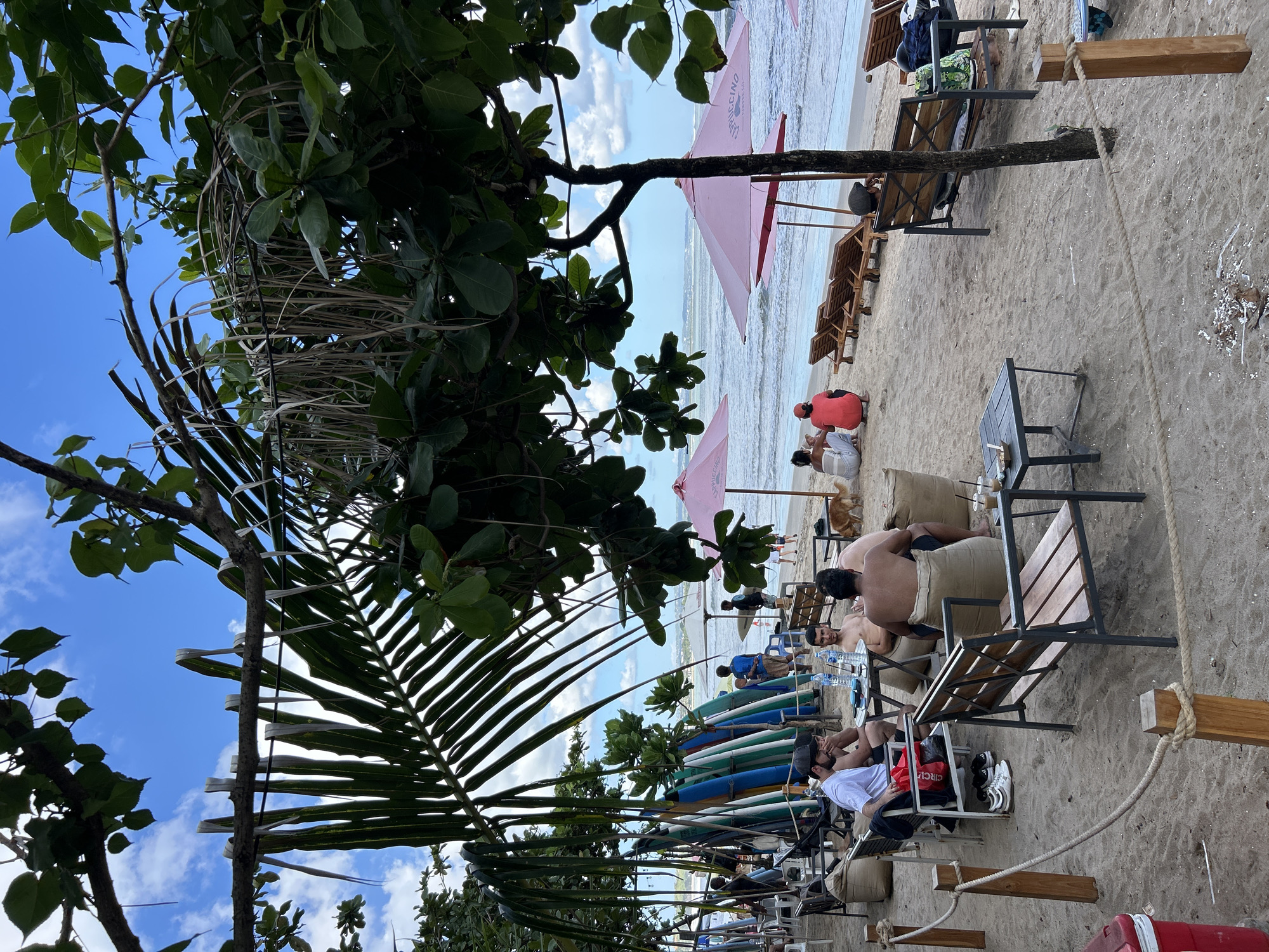 The image depicts a relaxed beach scene in Kuta, Indonesia, under a partly cloudy blue sky. In the foreground, several people are seated at tables and on beanbag chairs on a sandy beach. Some are engaged in conversation, while others are looking out towards the ocean. Surfboards are propped up in the background, suggesting a popular surfing destination. Two pink umbrellas are visible, providing shade for patrons. The ocean waves are gently rolling onto the shore. The overall atmosphere is one of leisure and recreation.