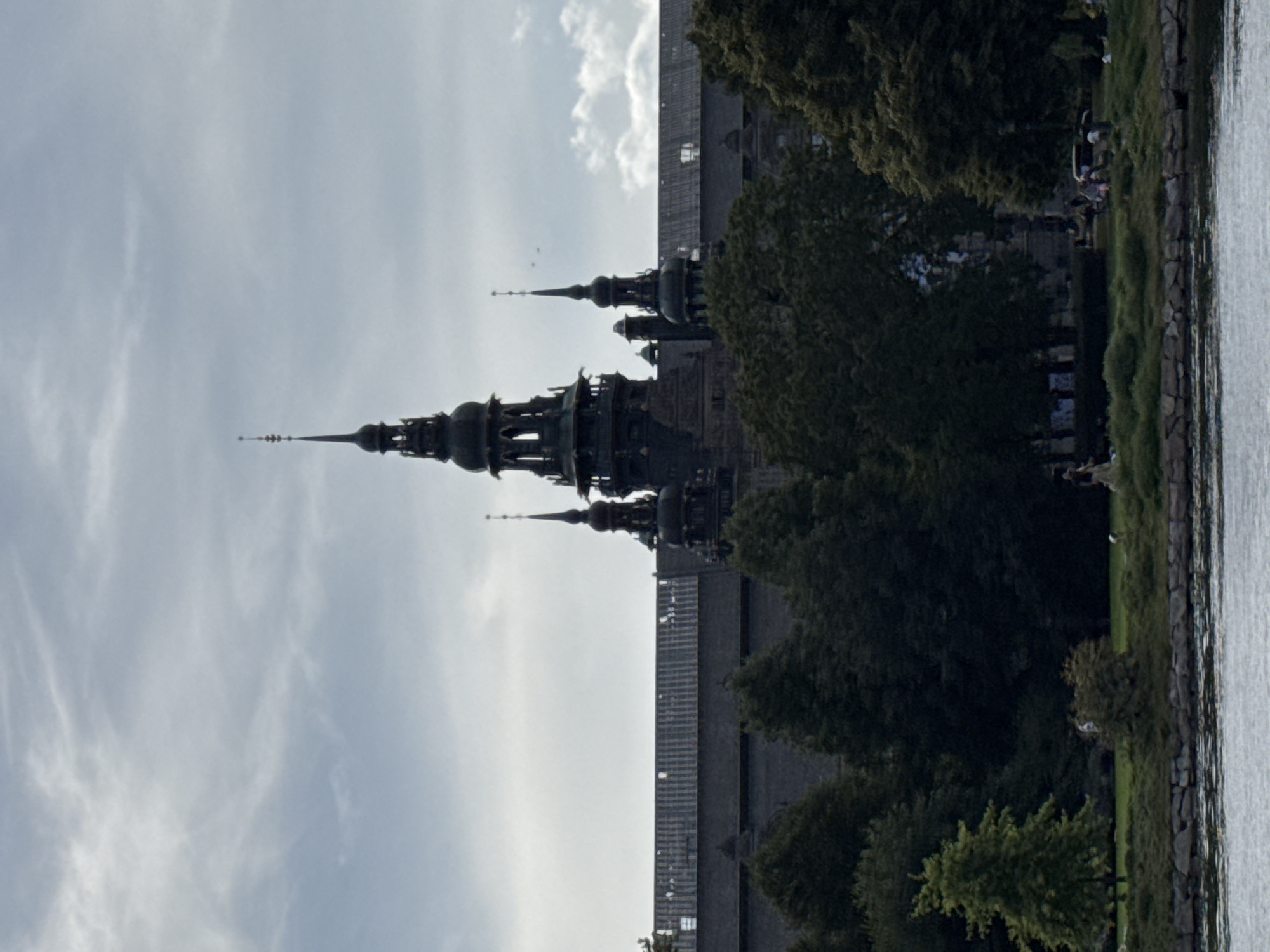 The image shows the Vasa Museum in Stockholm, Sweden, a large building with multiple towers and spires. The museum is situated on a waterfront with trees and green spaces in the foreground. Several people are visible in the park area, some appearing to be walking or relaxing. The sky is overcast with some clouds.
