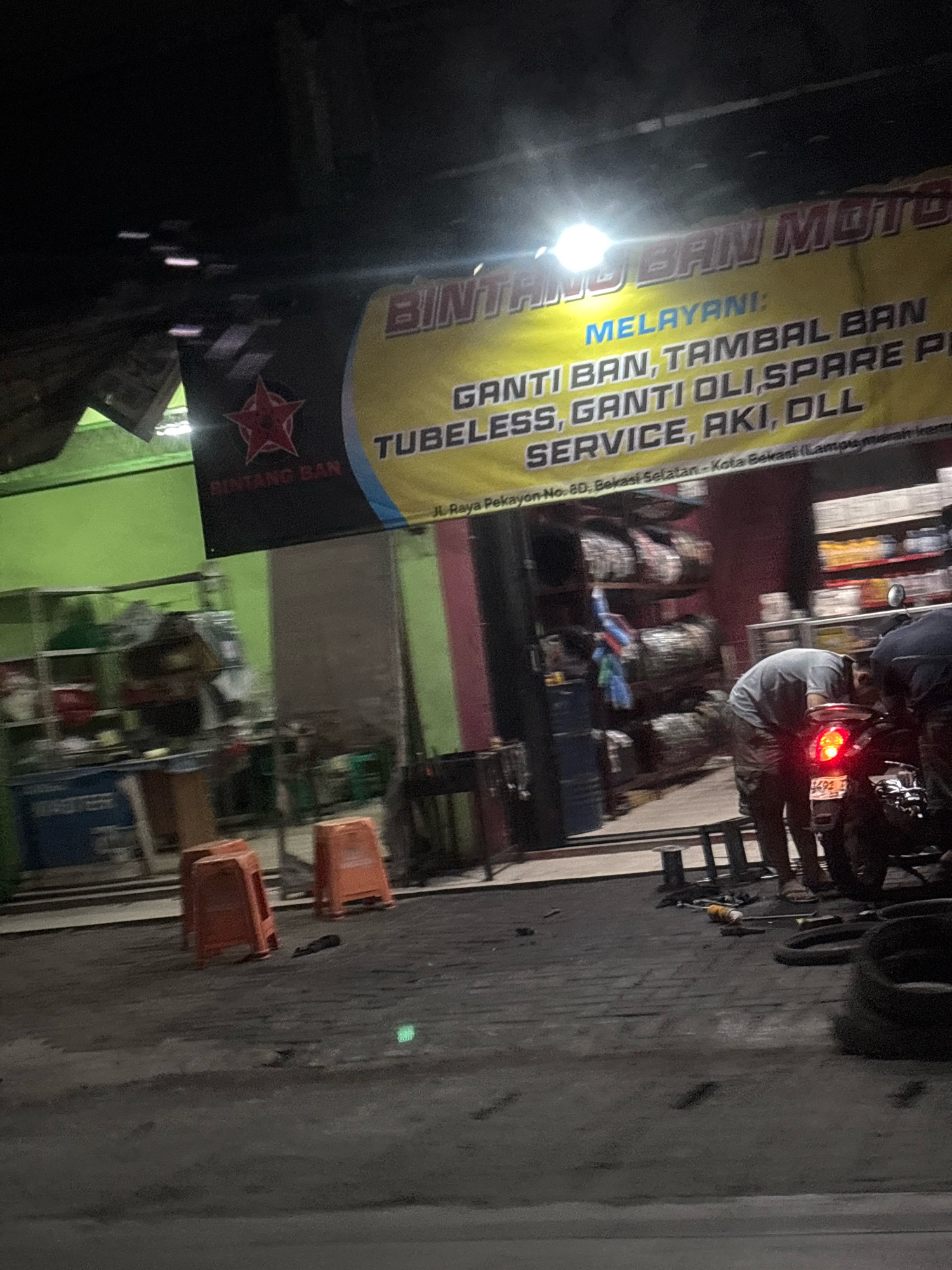The image shows a motorcycle repair shop at night in Bekasi, Indonesia. A large banner reads "BINTANG BAN MOTO" and lists services such as tire changes, patching, oil changes, spare parts, and general service. The shop appears to be located on Jl. Raya Pekayon No. 80, South Bekasi. Two individuals are working on a motorcycle, with one bending over the rear of the bike. Stacks of tires are visible in the background, along with shelves stocked with various items. Orange plastic stools are placed outside the shop. The scene is illuminated by a bright overhead light and the taillight of the motorcycle.