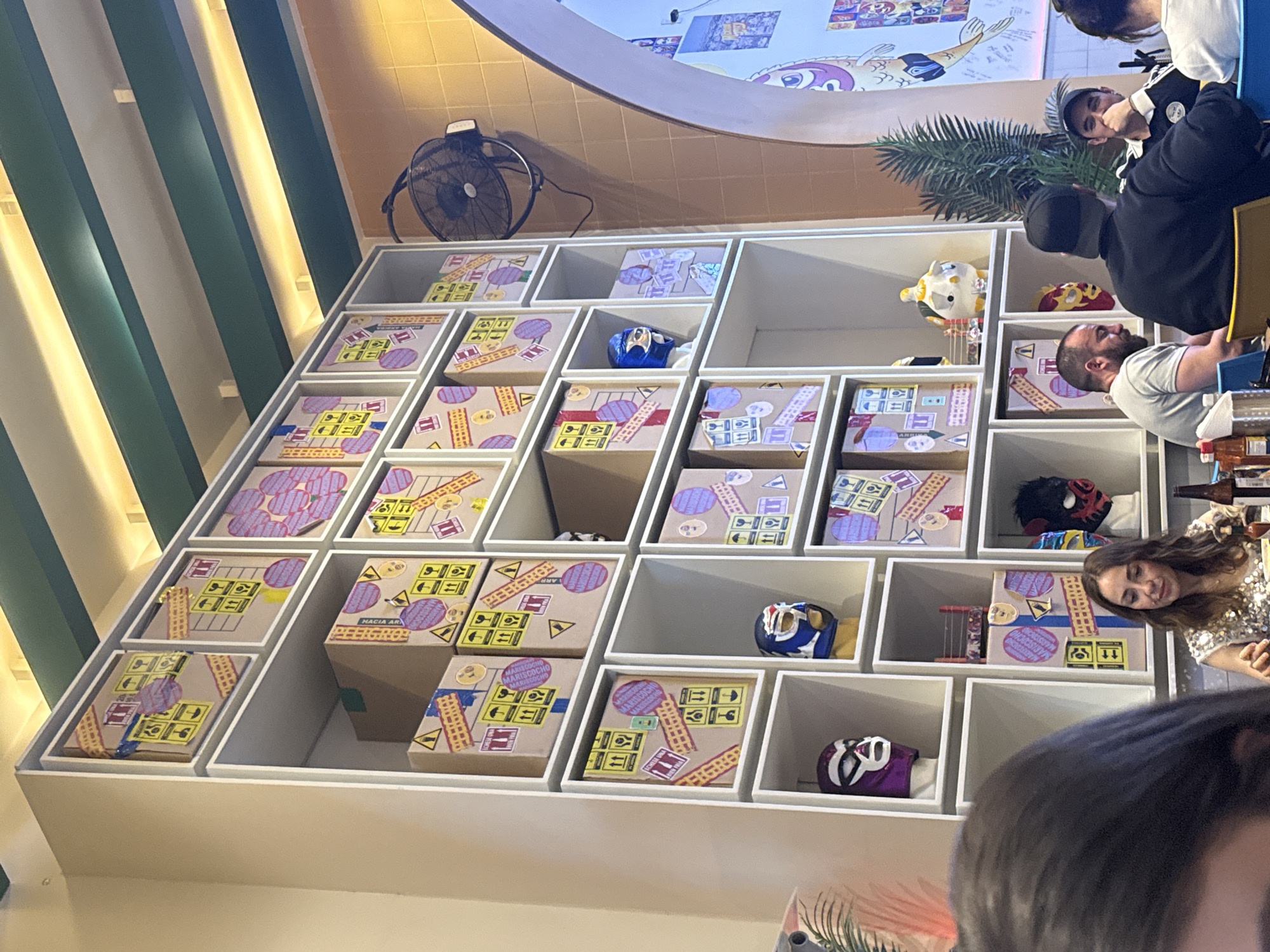 The image captures a unique decorative shelving unit. It has multiple square compartments, each holding an item or display piece. The shelving unit is partially visible. Some compartments contain boxes decorated with transportation-related symbols and writing in Spanish. Other compartments hold wrestling masks of various colors and designs.
People are seated at a table partially visible on the right side of the frame. They are engaged in conversation and interaction. The scene appears to be in a restaurant.
Additional details include a fan mounted on the wall above the shelving unit. Part of an artwork is visible on the wall. The scene takes place in Mexico City, Mexico, as implied by the cultural artifacts and the Spanish language visible on the boxes.