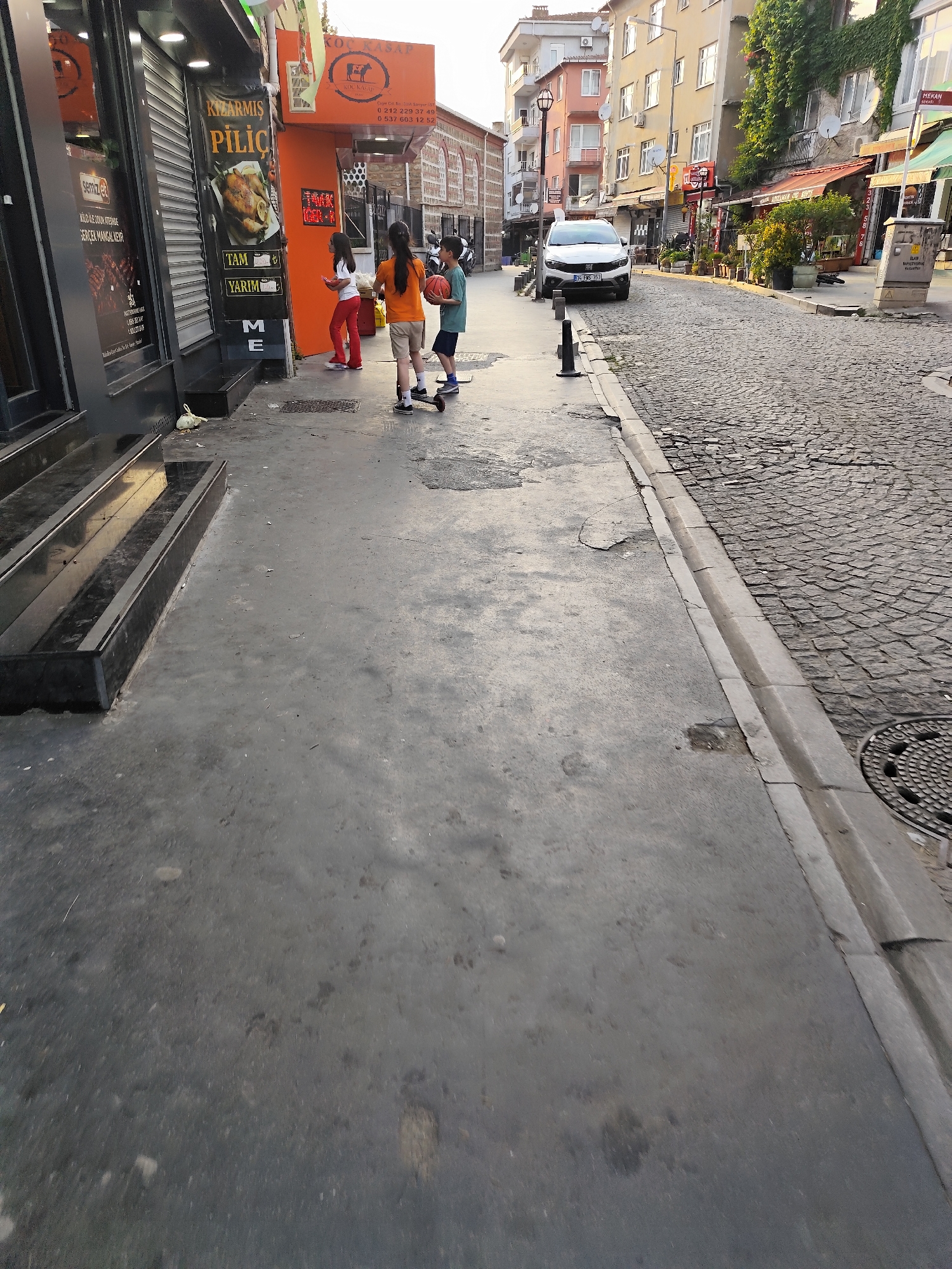 The image captures a vibrant street scene in Sarıyer, Turkey, during what appears to be late afternoon or early evening, characterized by warm, soft sunlight. The weather is clear and sunny.
In the foreground, a wide concrete sidewalk leads towards a cobblestone street. Three children are walking or playing on the sidewalk near the entrance of a shop. One girl, wearing red pants and a white top, stands near a storefront. Beside her, another girl in an orange top and shorts is positioned near a small, dark scooter. A boy in a blue top and shorts holds a basketball as he walks alongside the scooter.
To the left, a modern-looking shop facade features large display windows and black framing. Adjacent to it, an orange-colored establishment with a prominent sign for "KOÇ KASAP" (Koç Butcher) is visible. Further down the street, various buildings of different colors and styles line the road, some adorned with green ivy. A white Fiat car with the license plate "34 FMS 353" is parked further down the cobblestone street. Potted plants decorate the storefronts, and a utility box is visible on the right side of the street near a manhole cover.
Visible text includes:
- "KOÇ KASAP" (Koç Butcher) with phone numbers "0 212 229 37 49" and "0 537 603 12 52".
- A sign advertising "KIZARMIŞ PİLİÇ" (Roasted Chicken) with options "TAM" (Full) and "YARIM" (Half).
- Text related to a butcher shop or meat products: "SERNİZ ET", "KILO ILE DÖVÜN KTERNIZE GERÇEK MANGAL KEY" (likely referring to real barbecue pleasure with meat by the kilo).
- A street sign indicating "MEKAN" (Place/Venue).
- The license plate "34 FMS 353".
- Individual letters "M" and "E" on a lower sign.