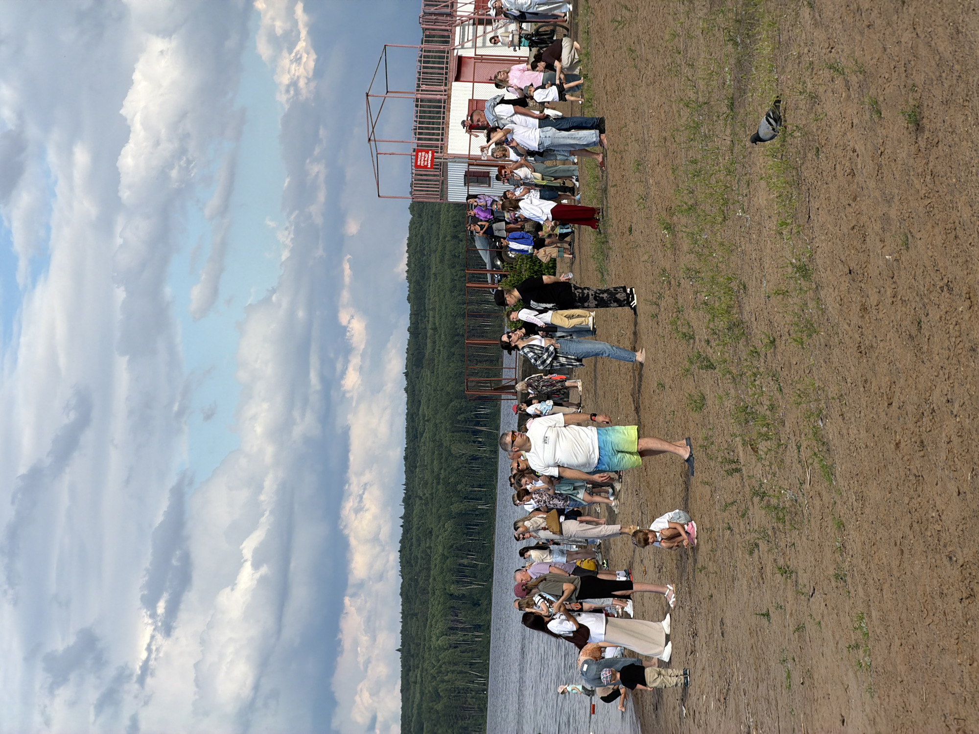 A group of people are gathered on a sandy embankment near a body of water, likely a lake or river, with a dense forest visible in the background. The sky is filled with dramatic, cloudy formations, suggesting a potentially dynamic weather system. In the distance, there is a multi-tiered structure that resembles bleachers or a viewing platform, with some people positioned on or near it.
The people are of various ages and are casually dressed, suggesting an outdoor gathering or event. Some are standing in a line, while others are interacting in smaller groups or observing the scene. One person is wearing colorful swim shorts, and another is holding what appears to be a phone. There's a pigeon on the ground in the foreground. The overall atmosphere seems relaxed and informal, possibly a public event or a casual outing by the water. The lighting suggests it is daytime, likely afternoon or late morning, given the bright sky and shadows. A sign on the structure in the background contains Cyrillic text.