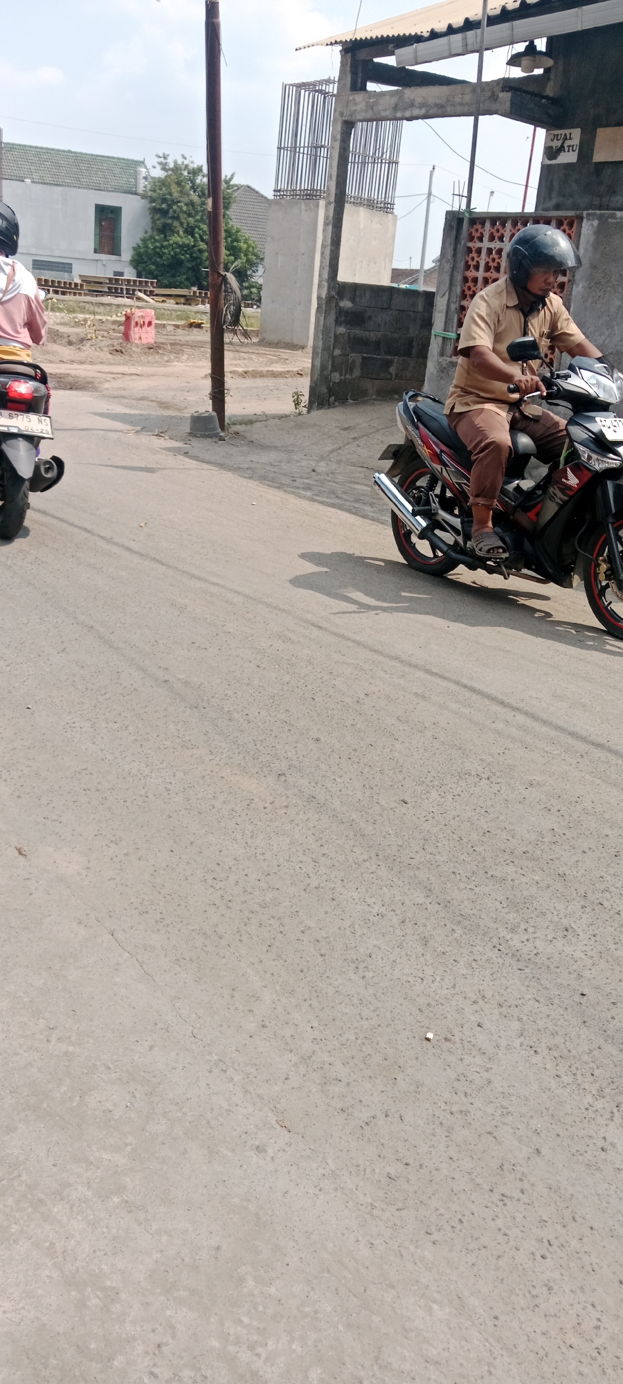 In Purwomartani, Indonesia, a man in a tan shirt and brown pants is riding a black motorcycle. He is wearing a black helmet. The motorcycle has red accents and a silver exhaust pipe. The man is on a paved road, with construction in the background. The construction area has concrete pillars and rebar, as well as a partially built structure with a corrugated metal roof. A white building with a green roof is visible in the distance, along with some trees. Another motorcycle, mostly out of frame, is to the left. The road is worn and appears to be in a residential or developing area. The sky is clear and blue.