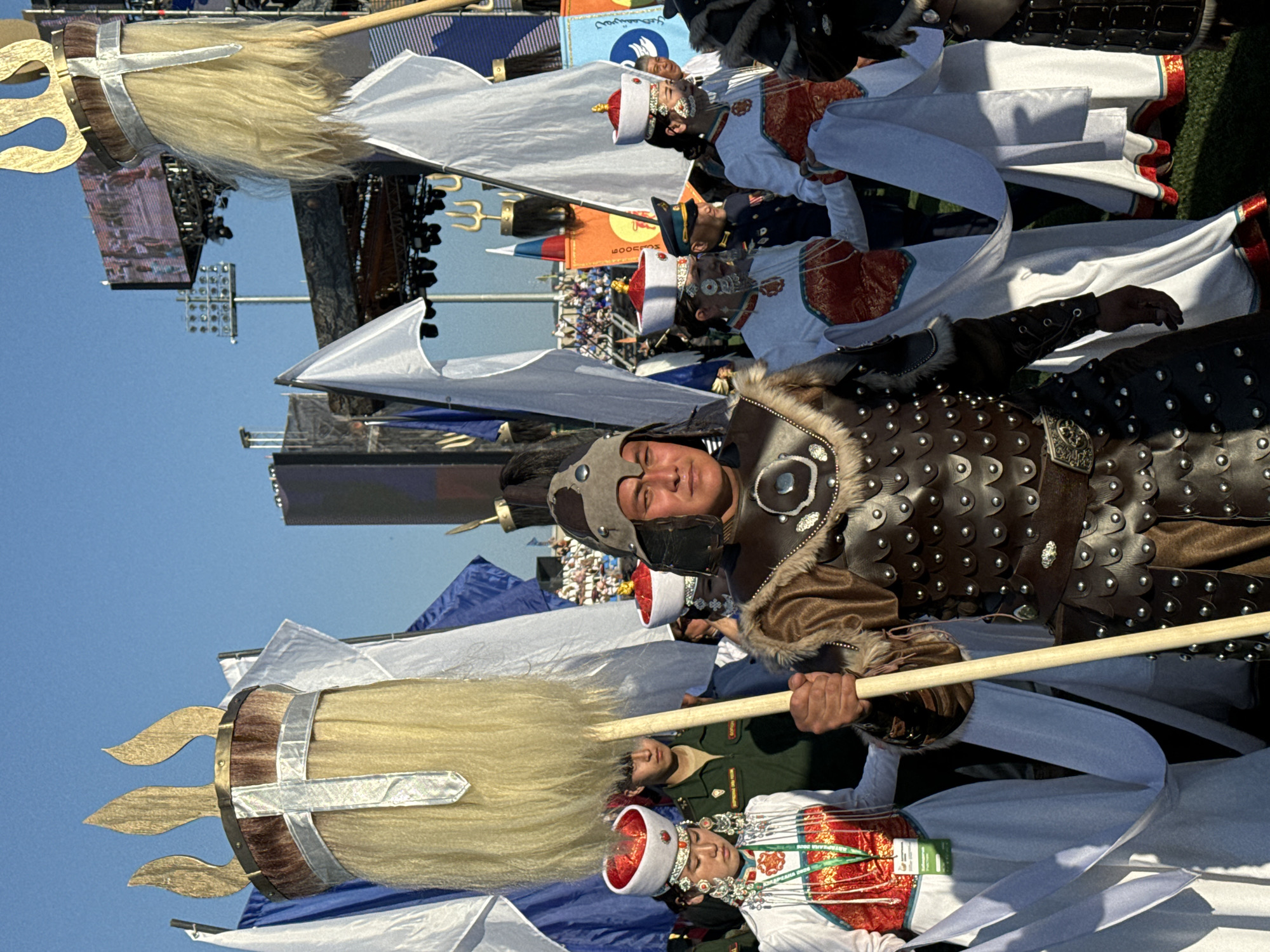 This media file shows a procession or parade in Ulan-Ude, Russia. The subjects are primarily people dressed in traditional or ceremonial attire, possibly representing historical or cultural figures. They are holding large, ornate poles topped with blonde hair and golden trident-like ornaments. Some individuals wear armor-like vests, helmets, and detailed hats with red and white accents, adorned with intricate jewelry. Others are in more formal, perhaps military-style uniforms.
The scene appears to be outdoors, with a clear blue sky suggesting a sunny day. In the background, there are stages, lighting rigs, and what looks like a crowd of spectators, indicating a public event. The ground appears to be covered in grass in some areas. The overall atmosphere seems festive and ceremonial, with participants displaying pride in their heritage through their elaborate costumes. There are flags visible, one with Cyrillic text. The faces of the participants show a range of expressions, but many appear calm and focused, possibly indicative of the solemnity of the occasion or their role in the procession. The lighting suggests it is daytime.
