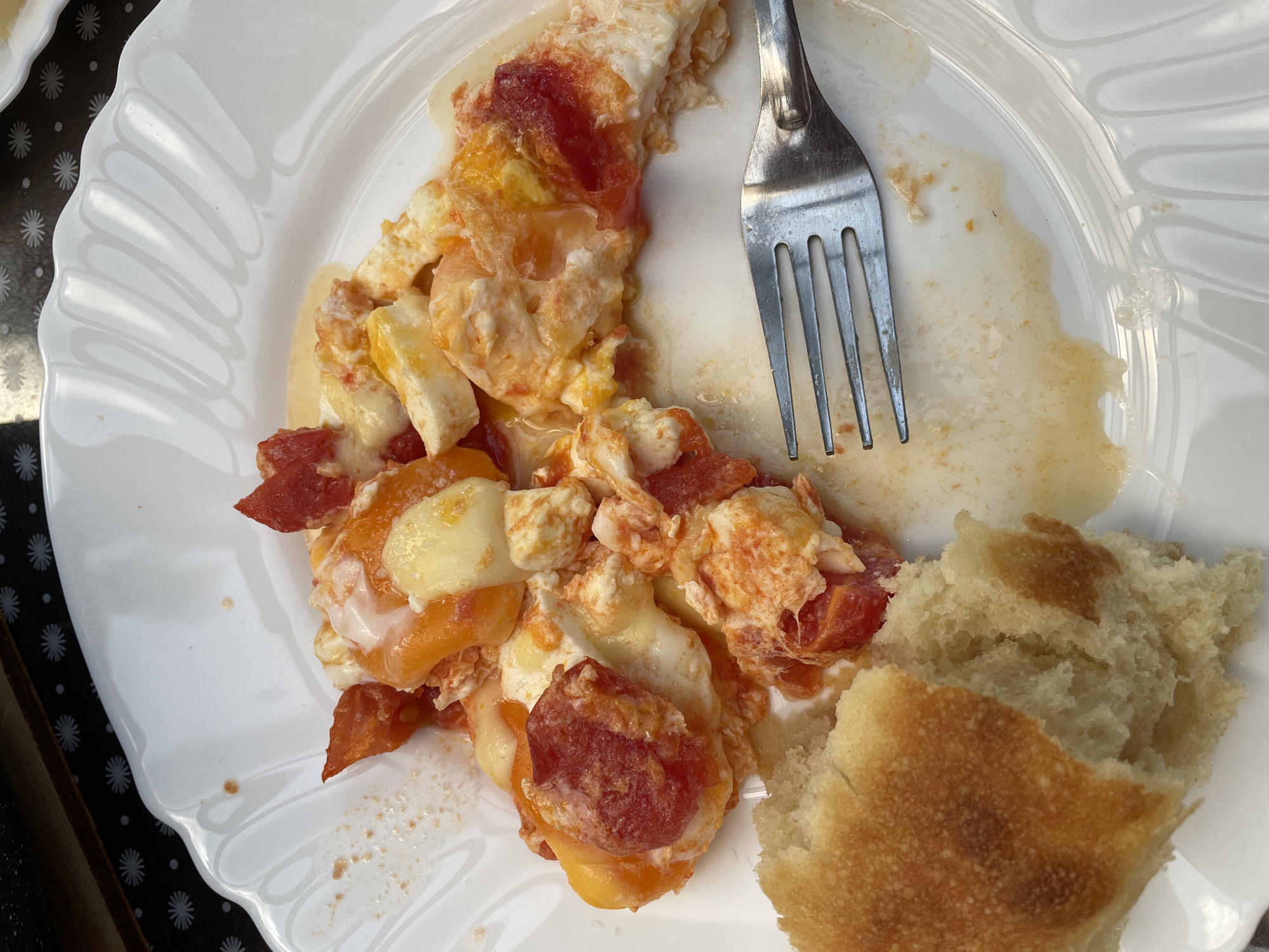A close-up, overhead shot shows a white, fluted plate holding a portion of a breakfast dish. The dish appears to be scrambled eggs with diced tomatoes and possibly sausage. A piece of bread is on the left side of the plate. A fork is positioned on the right side of the plate. The plate is set on a dark placemat with a white starburst pattern. The lighting suggests the image was taken outdoors during the day. The scene is an unknown location in an unknown city.