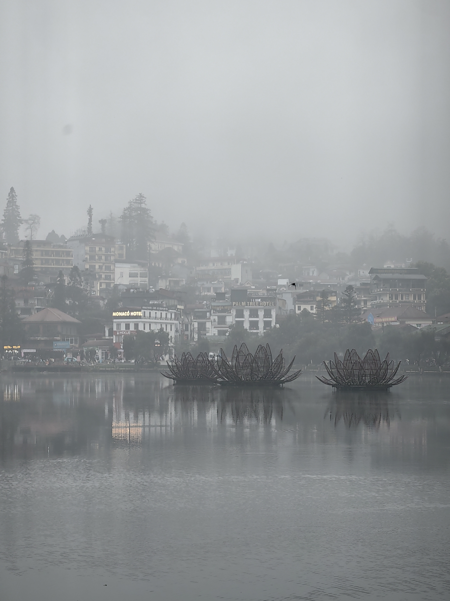 The image depicts a foggy scene of Sa Pa Lake in Sa Pa, Vietnam, with the town's urban landscape rising on the far bank. The environment is dominated by thick, gray fog, obscuring the sky and distant hills, and muting the color palette.
In the foreground, a calm body of water occupies the lower half of the frame, exhibiting subtle ripples across its surface and reflecting the lights and structures from the opposite shore. Three large, dark, open-weave structures, resembling stylized lotus flowers made of reeds or wood, are positioned on the water, slightly to the right of the center, reflecting in the water below them.
The midground features the densely packed architecture of Sa Pa town along the lake's edge and climbing uphill. Buildings are multi-story, exhibiting various architectural styles and light-colored facades. Notable illuminated signage includes "MONACO HOTEL" and "MIXUE" in the center, and a partial "PALM TREE HOTEL" on the left. Scattered figures, likely pedestrians, are faintly visible along the waterfront promenade. Interspersed among the buildings and on the slopes above are dark evergreen trees, partially obscured by the fog. A small, dark bird is captured in flight above the lake, against the uniform gray background of the fog.
The overall scene suggests a cool, damp atmosphere with limited visibility due to the pervasive fog, possibly during dusk or nighttime given the illuminated signs. No direct interactions between individuals are discernible.
