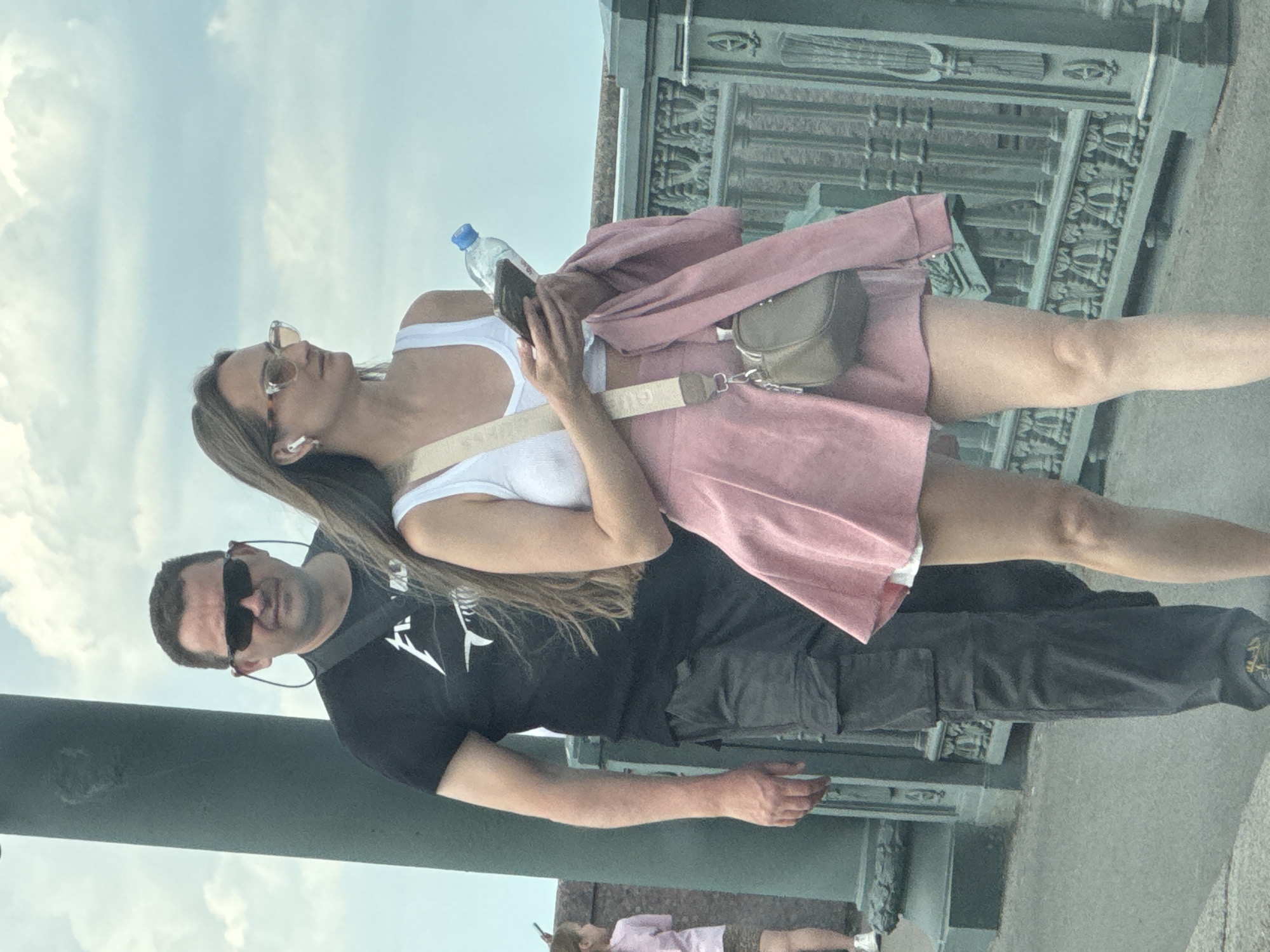 The image captures two individuals, a woman and a man, standing outdoors on what appears to be a bridge or elevated walkway in Saint Petersburg, Russia.
The woman, appearing to be a young adult with long brown hair, is positioned on the left. She wears sunglasses, a white tank top, a light pink jacket, and a matching pink mini skirt. She has white earbuds in her ears and carries a small olive-green cross-body bag, partially revealing "GUE" (likely "GUESS") on its strap. In her hands, she holds a smartphone and a clear plastic water bottle. She is looking upwards and slightly to her left, possibly at the sky or something above her.
Beside her, to the right, stands a man, likely in his 30s or 40s, wearing black sunglasses, a black t-shirt with white graphics (resembling a stylized skeletal fish or bird), and dark grey cargo pants with a small, indistinct logo on the lower right leg. He looks forward with a neutral expression. In the distant background, to the lower right of the man, a small, partially obscured figure in a pink top is visible.
The scene is set on a bright, cloudy day, with a pale blue sky and white clouds overhead. A distinctive, ornate dark green/grey metal railing or architectural structure, characteristic of Saint Petersburg's bridges and embankments, forms a backdrop to their left. The two individuals appear to be enjoying a casual day out.