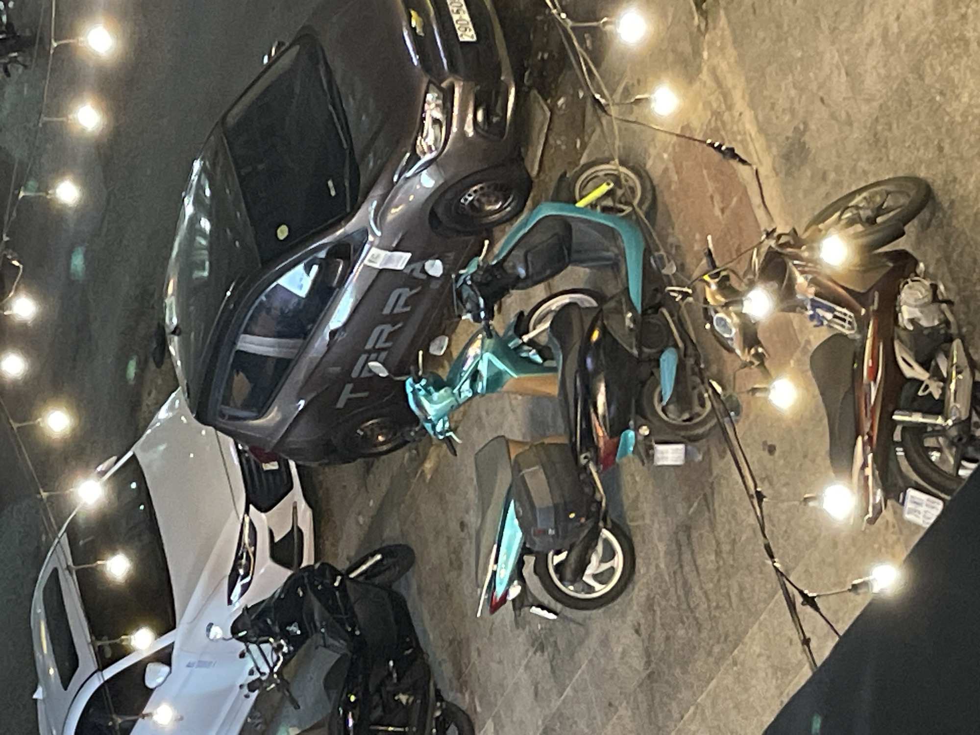 This night-time image, captured in what appears to be a street or parking area in Bắc Nha Trang, Vietnam, features several parked vehicles illuminated by string lights. The ground is paved concrete.
In the center-left, a dark grey Nissan Terra SUV is prominently parked, with "TERRA" clearly visible on its side. A partially visible license plate reading "29D-505..." is affixed to its front. A white sedan is parked behind it to the far left.
Closer to the foreground and on the right side of the frame, multiple motorbikes and scooters are parked. A distinctive turquoise/teal scooter occupies the central foreground, while a brown or maroon scooter is parked further to the right. Another dark-colored scooter is partially visible in the bottom-left corner.
The scene is lit by numerous bright, warm-toned string lights strung overhead, creating glows and reflections across the vehicles and ground, indicating it is dark outside. No people are visible, and there is no active event taking place, just a collection of parked vehicles.
