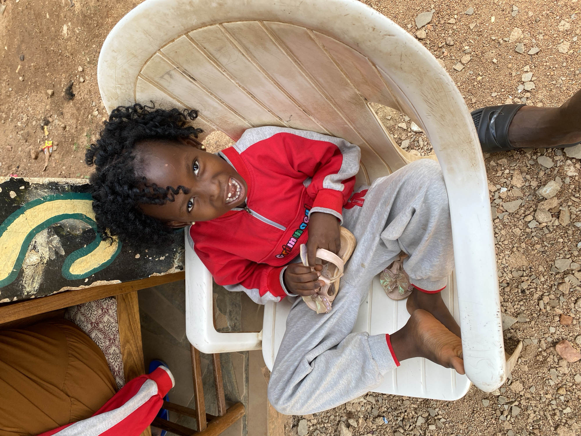 A young child with dark skin and curly black hair is reclining in a white plastic chair outdoors. The child wears a red and gray tracksuit and is holding a pair of light pink sandals in their hands. The child is smiling and looking upwards. The ground around the chair is dirt and small rocks. Adjacent to the chair is a wooden table. An adult's leg and foot wearing a black sandal is visible on the right side of the image. The location is Kuru Karama, Nigeria.