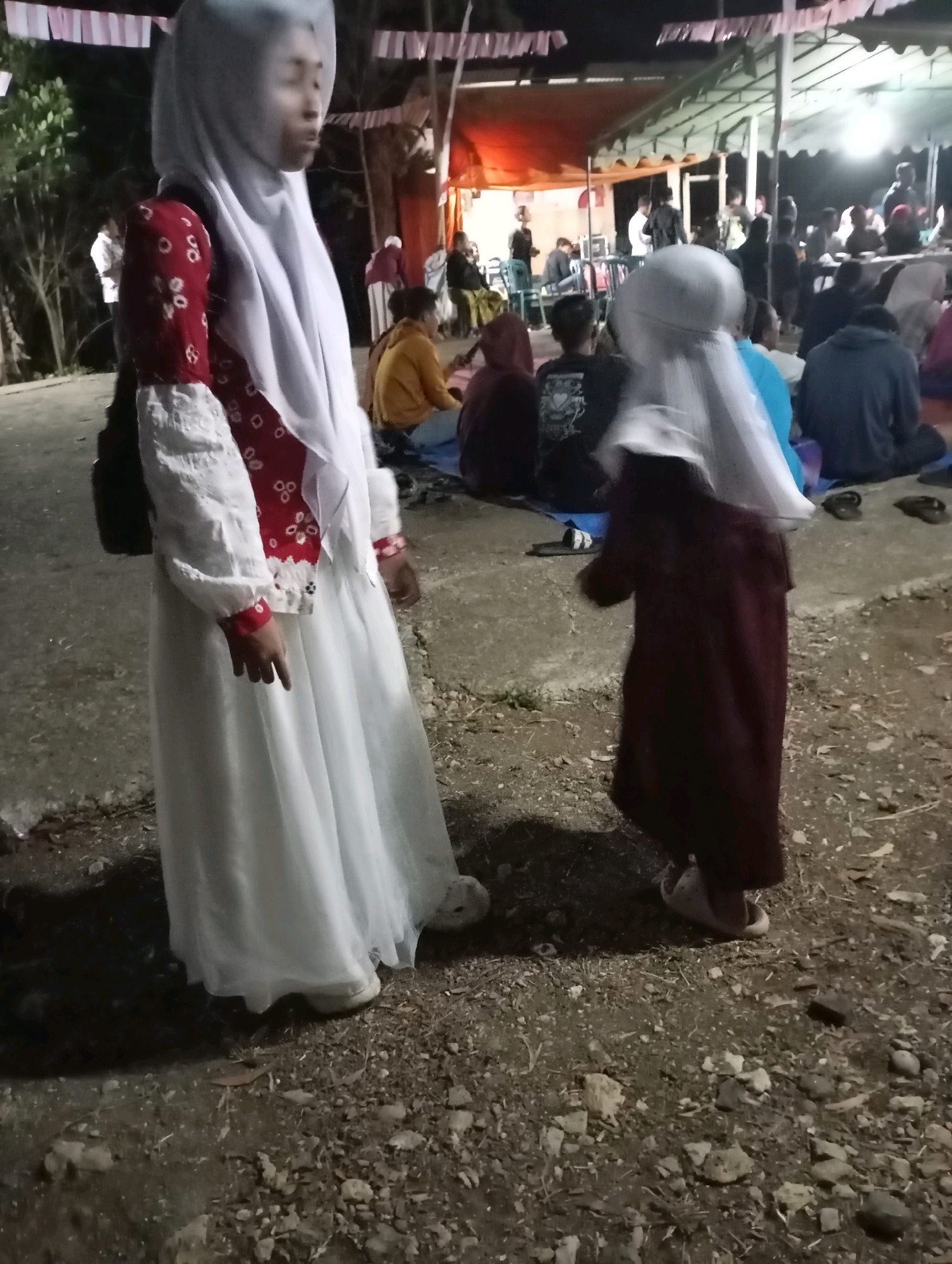 Two individuals, seemingly children or young women, are standing on a dirt ground. The individual on the left is wearing a white hijab and a white long-sleeved dress with red accents. The individual on the right is wearing a dark-colored hijab and a dark-colored dress. In the background, there are several other people, some seated and some standing, under a tent-like structure. The scene appears to be an outdoor gathering, possibly a community event or celebration, in a location that may be Wonogondo, Indonesia, given the context provided. The lighting suggests it is nighttime or dusk.