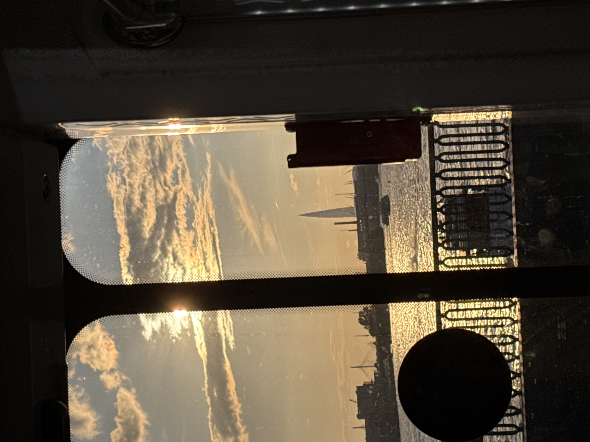 This image depicts a reflection in a window, likely of a vehicle, showing a sunset over a body of water with a city skyline in the distance. The sky is filled with dramatic clouds illuminated by the setting sun, creating warm orange and yellow hues. In the middle ground, a sailboat is visible, and beyond it, the silhouette of buildings and structures of a city can be seen. The scene suggests a peaceful evening in a coastal city, possibly Saint Petersburg, given the mention of Russia in the context. The reflection also shows parts of the interior of the vehicle, including a dark object which could be a headrest or a part of the car's interior, and a coiled wire structure which appears to be part of the window mechanism or an accessory. The overall mood is serene and contemplative, enhanced by the beautiful natural light. There is no visible text in the image.