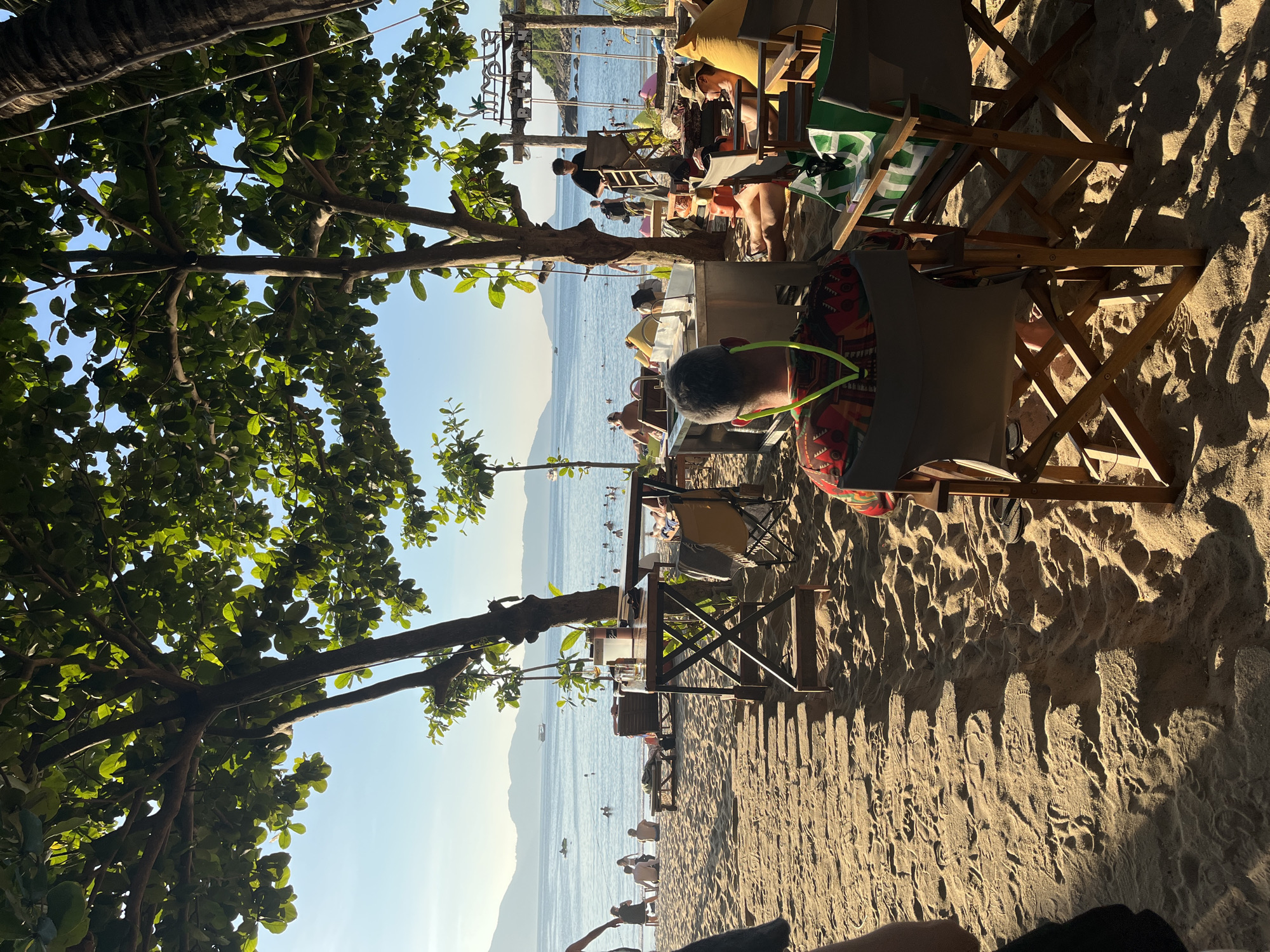 This image captures a vibrant beach scene, likely in Đà Nẵng, Vietnam, under a bright blue sky. The perspective is from a shaded area beneath lush green trees, looking out towards the ocean.
In the foreground, several people are visible. One person, seen from behind, wears a colorful, patterned shirt and what appears to be bright lime green headphones or a lanyard, seated at a wooden table on the sandy beach. Adjacent to them are wooden beach chairs and tables, some with drinks. Long, distinct shadows of the chairs and trees stretch across the coarse, light brown sand, indicating a late afternoon or early evening setting with warm, golden sunlight. Footprints are visible in the sand, suggesting recent activity.
Further into the mid-ground, more people are scattered along the beach and in the calm, turquoise ocean. Some are seated at additional tables and chairs set up closer to the water's edge, while others are swimming or wading in the shallows. Small boats are dotted across the water. In the distance, gentle mountains rise from the far side of the bay, completing the picturesque coastal landscape.
The overall atmosphere suggests relaxation and leisure. While no clear text is decipherable on signs or objects, the scene conveys a typical tropical beach resort or cafe environment.