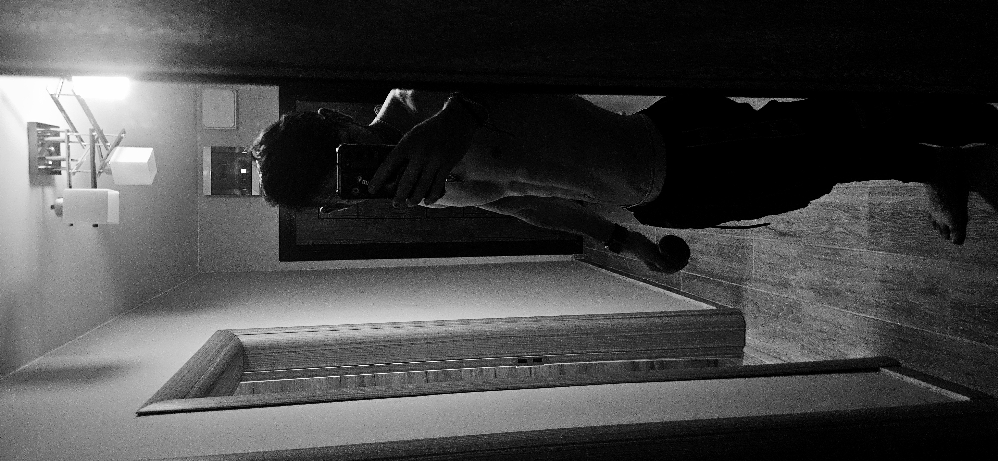 A monochrome image depicts a man in a mirror reflection, likely taking a photograph of himself. The man is shirtless, wearing dark bottoms, and holding a smartphone. He has short, dark hair and appears to be in his bathroom. The bathroom contains a light fixture with multiple lamps, a framed mirror, and tiled flooring. A second doorway or entryway with wood trim is also visible in the lower portion of the image. It is not possible to determine whether the location is in the city of Plovdiv, Bulgaria.