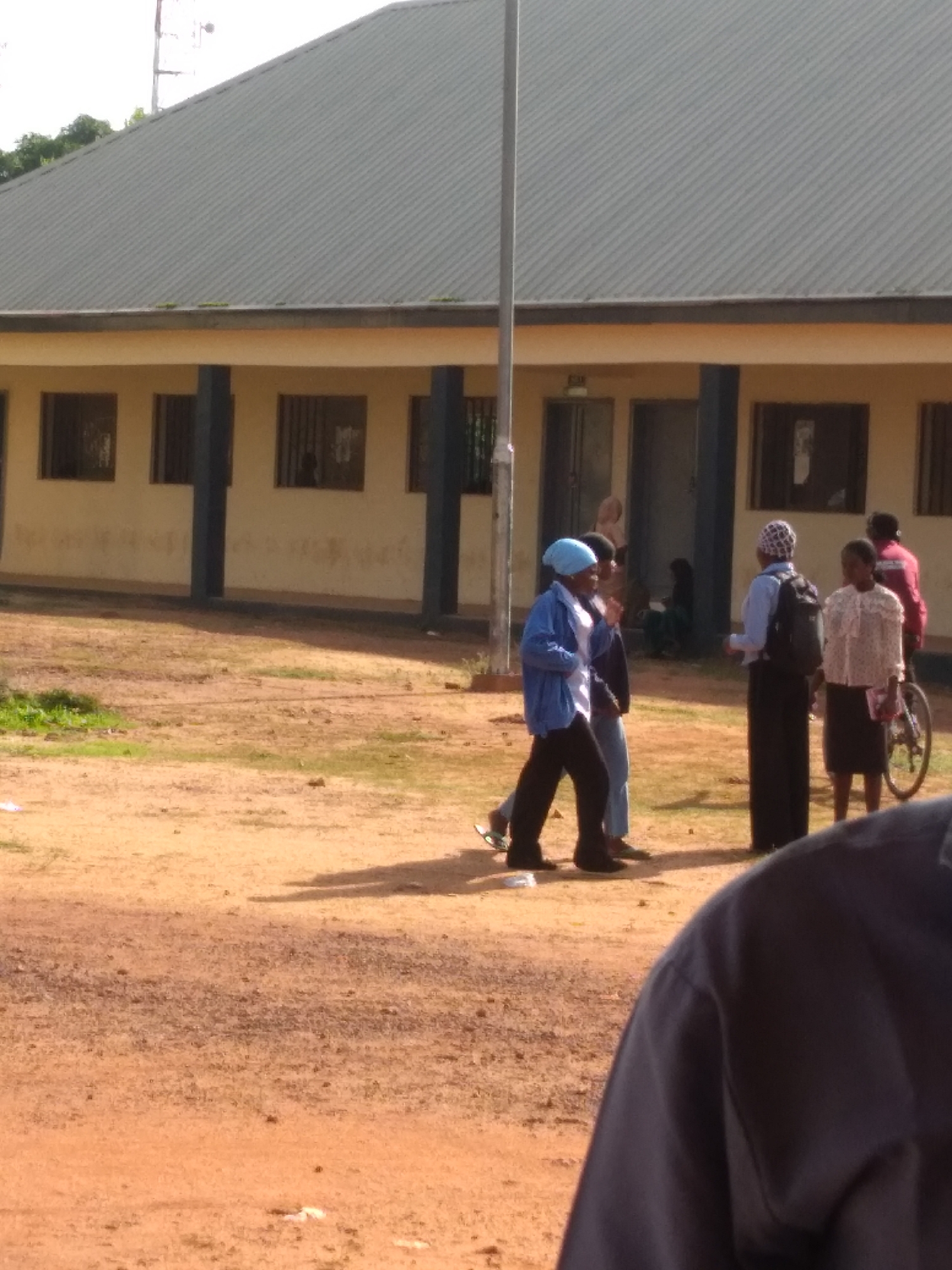 A group of people are gathered outdoors in front of a light-colored, single-story building with a dark, corrugated metal roof. The building has several windows with dark bars and dark vertical pillars supporting an overhang. The ground is a dusty, reddish-brown dirt with patches of dry grass. In the foreground, a person wearing a dark blue shirt is partially visible, obscuring the lower portion of the image. To the left, a young person in a light blue jacket and light blue headscarf is walking, with another person in dark pants to their right. Further back, a person in a patterned headscarf and dark clothes stands near a backpack, facing another person in a patterned top and dark skirt. A bicycle with a person in a red jacket standing behind it is visible on the far right. In the background, some trees are visible to the left of the building, and a tall, thin structure, possibly a radio tower, is present in the upper left corner. The scene appears to be in an open area, perhaps a school or community center, under a bright sky, consistent with the environment in Jos, Nigeria.