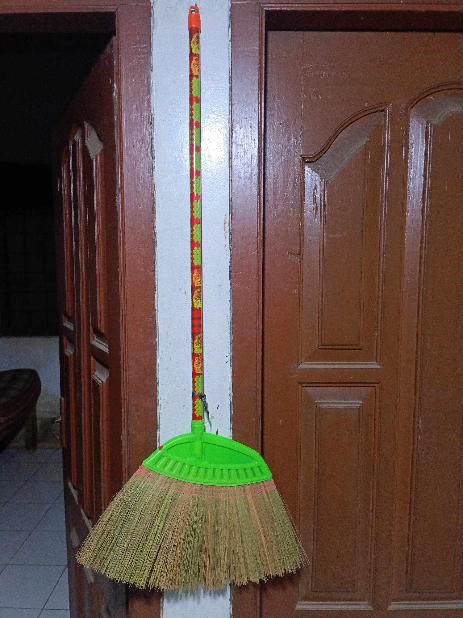A broom with a green plastic head and a red, green, and yellow patterned handle hangs from a hook on a white wall. The broom is positioned between a dark brown wooden door on the left and another dark brown wooden door on the right. The door on the left is slightly ajar, revealing a dark interior. The door on the right has multiple recessed panels. The floor visible in the background appears to be tiled. The image was taken in Jenu, Indonesia.