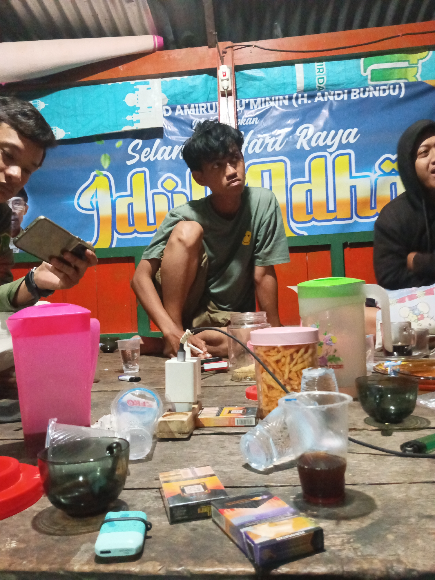 The image depicts an indoor gathering with at least three young men seated around a wooden table. The men appear to be of Indonesian descent. One man on the left is looking at his phone, while another in the center is looking downwards. The third man on the right is partially obscured. The table is cluttered with various objects including a pink pitcher, a green-lidded pitcher, a glass jar filled with snacks, cigarette packs, a small light blue device, a white electronic device, and several used plastic cups, some containing a dark liquid. In the background, a blue banner with white mosque imagery and text is visible, along with a section of a corrugated metal roof. The scene is lit by artificial light, creating shadows. The presence of a banner with what appears to be Indonesian text suggests the location is likely within Indonesia. While the specific location within Tumampua is not directly indicated, the general setting suggests a casual, possibly informal social gathering.