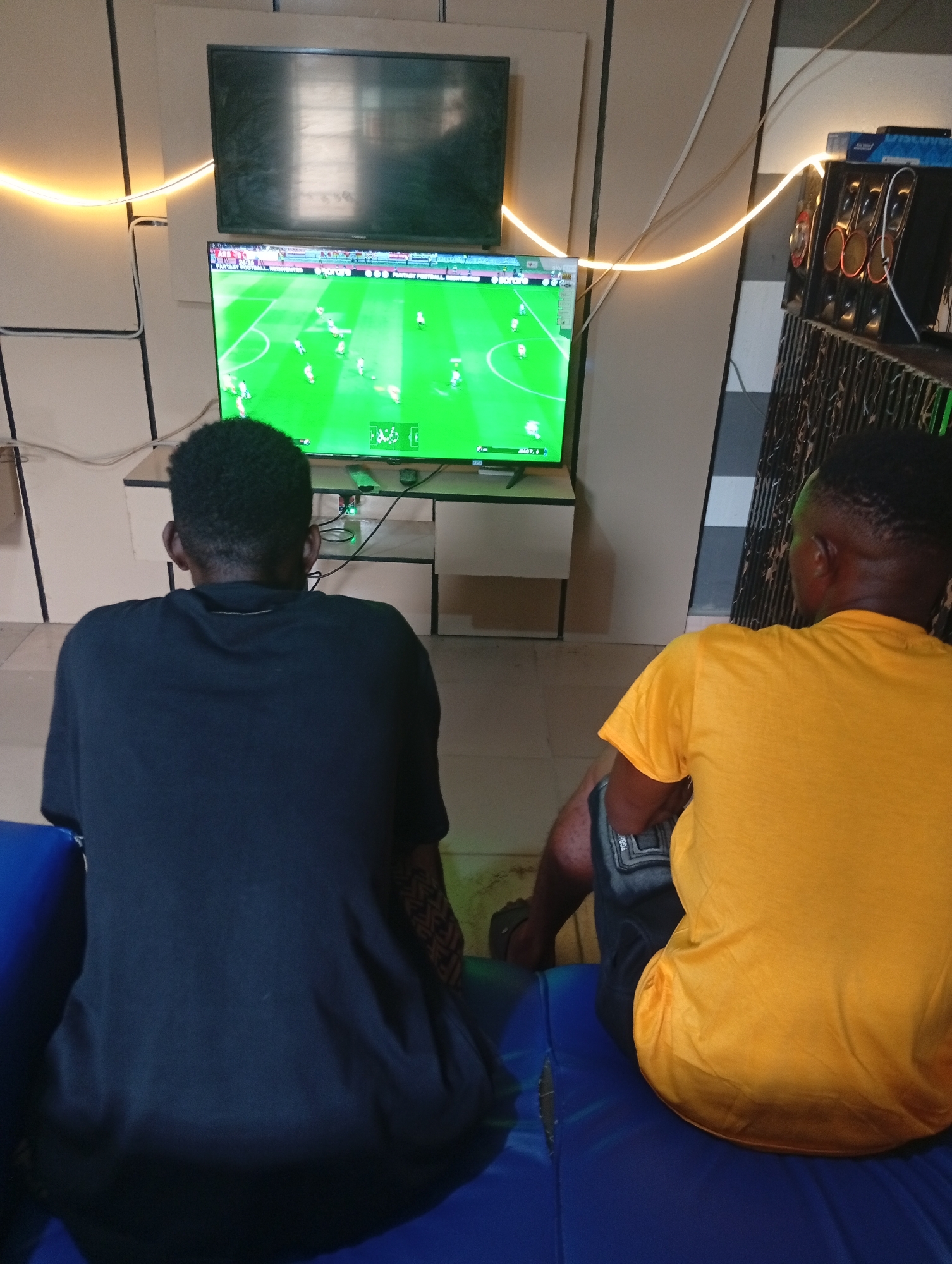 Two men are depicted from behind, deeply engaged in playing a soccer video game on a large television screen in an indoor setting in Adankolo, Nigeria. They are seated side-by-side on blue cushioned benches or seating, their backs to the viewer, indicating their focus on the screen.
The man on the left wears a black t-shirt, while the man on the right is dressed in a vibrant yellow t-shirt and dark shorts. The main television screen displays a bright green soccer field with numerous animated players actively involved in a match. Partially visible text on the game interface at the top left reads "FANTASY FOOTBALL." Above this active screen, a second, larger television is mounted, which appears to be off or displaying a dark image.
The room features a backdrop of light-colored wall panels with dark vertical divisions. A distinct glowing yellow light strip curves along the wall, providing ambient lighting and a decorative touch. To the right of the setup, a stack of audio speakers or entertainment equipment is visible on a dark shelf, connected by several wires. A simple, light-colored stand supports the main television. The overall scene suggests a casual gaming environment, possibly a dedicated gaming room or a local gaming lounge, designed for immersive entertainment. The artificial lighting prevents determination of the exact time of day.