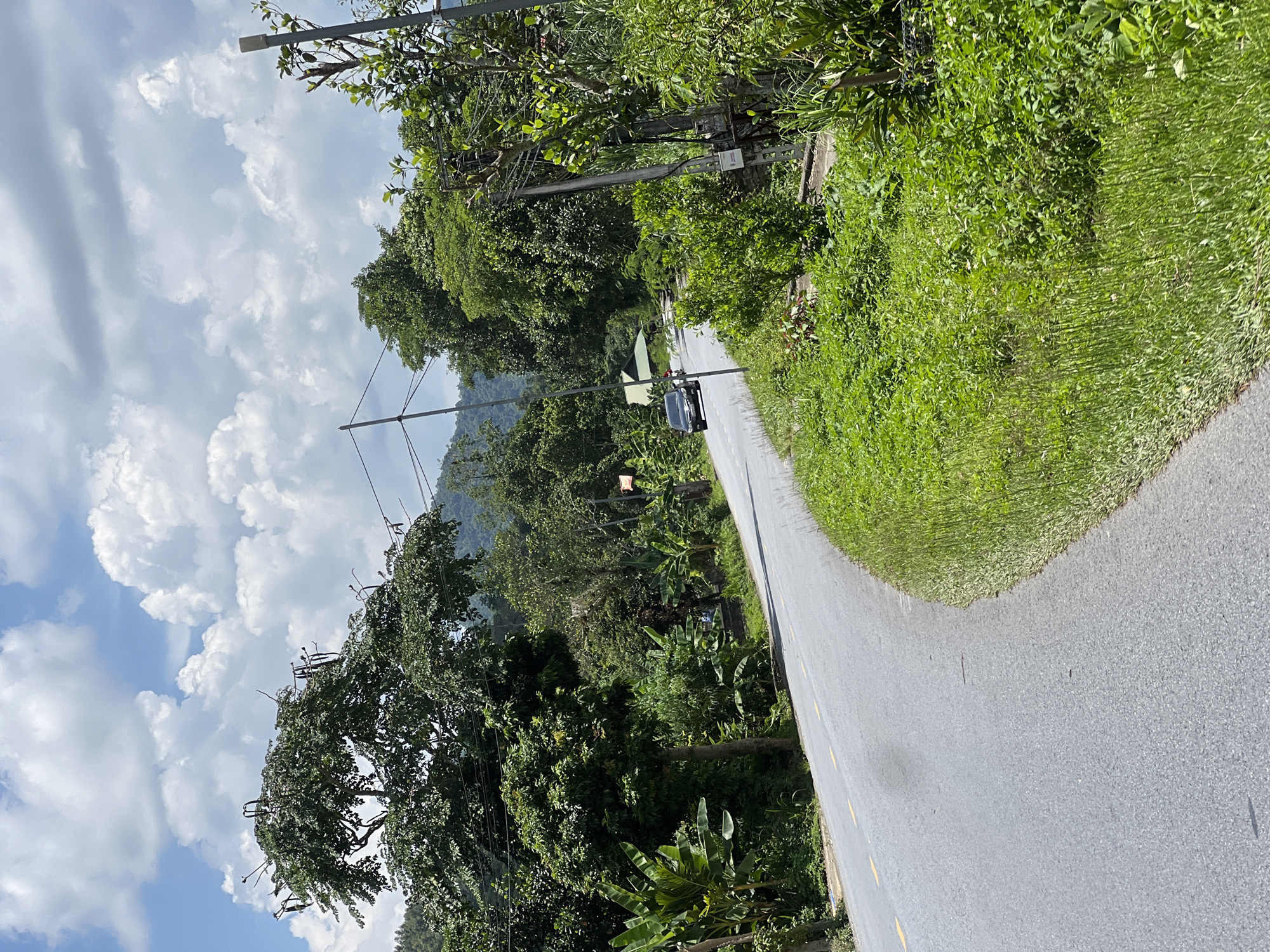 The image depicts a curving asphalt road with yellow lane markings, bordered by lush green vegetation on both sides. A dark-colored car is visible driving away from the viewer on the left side of the road. In the background, a hilly landscape is covered with dense trees, and a few buildings with green roofs are nestled amongst the foliage. The sky above is partly cloudy with white cumulus clouds against a blue backdrop. The scene appears to be a rural or semi-rural area. Based on the provided context, this scene is located in Thuận Hòa Commune, Vietnam.