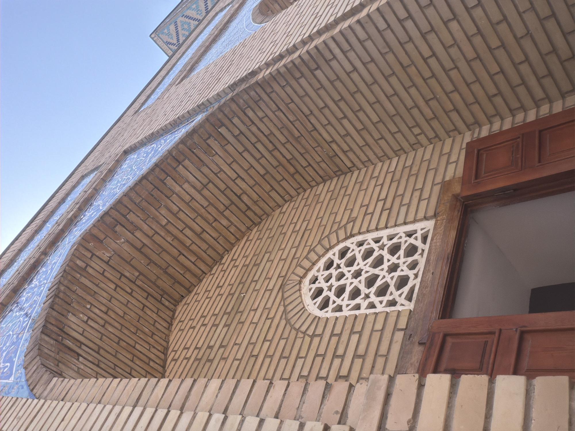 The image shows a low-angle view of the exterior of a brick building. The building features decorative blue tiles with white patterns along the upper facade and an arched opening with a white, geometrically patterned lattice window. Below the window, a dark red wooden door is ajar, revealing a dark interior. The brickwork is light tan with darker mortar. The sky is a clear light blue. The architectural style suggests it is likely a historical site in Zangiata, Uzbekistan, possibly a mausoleum or mosque, given the common decorative elements and materials found in the region. No people are visible in the image.