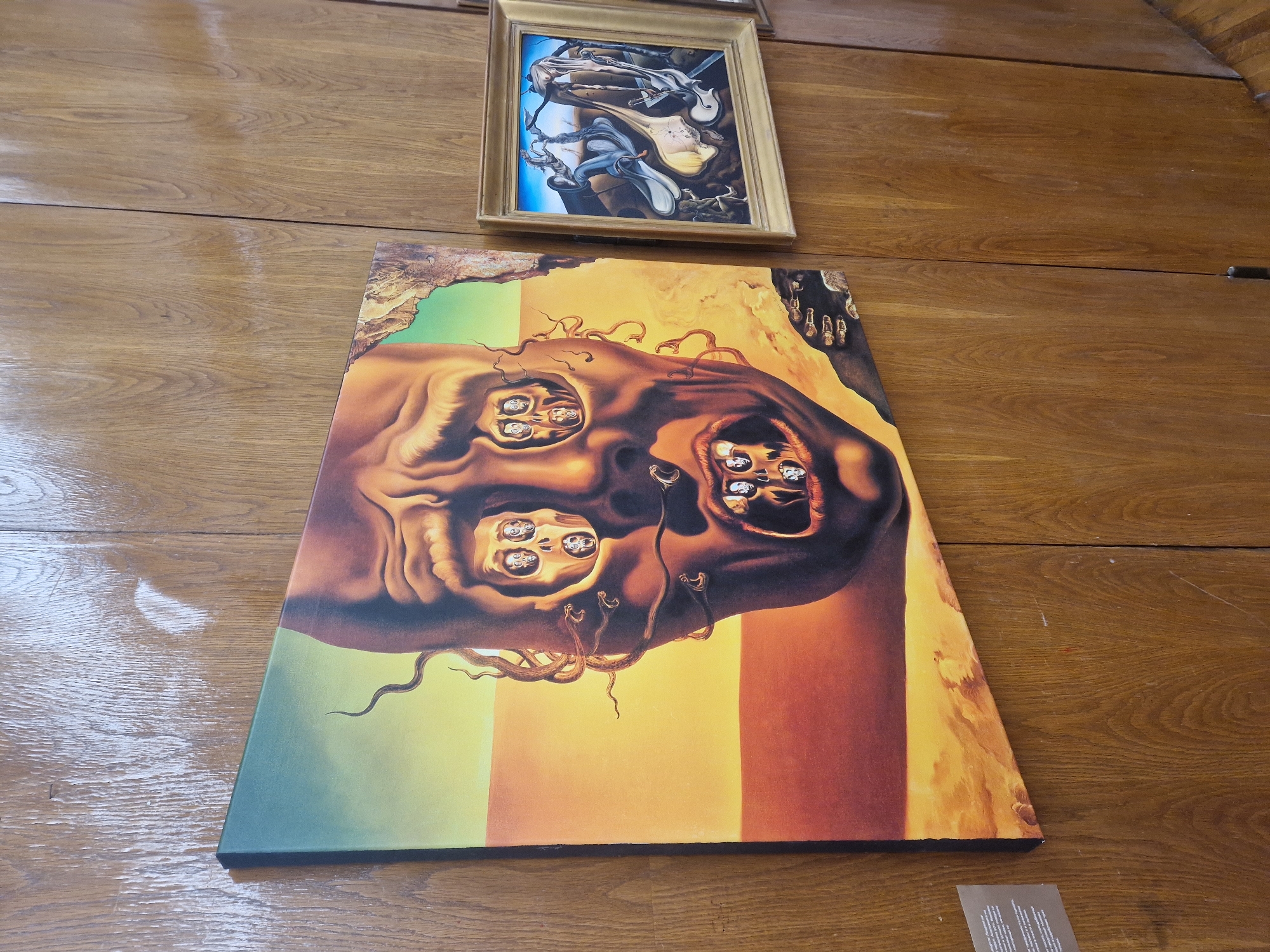 A view of a wooden table with two paintings on it. One is rectangular and depicts a skull with several faces in its eye sockets. The second painting is smaller, framed, and features abstract figures in a surreal composition. The location is Unknown, Unknown.