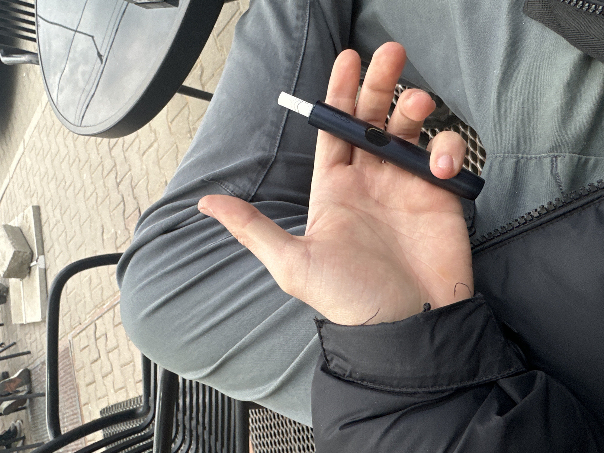 A person is seated at an outdoor cafe, likely in Plovdiv, Bulgaria, although without more context this cannot be definitively stated. The person is holding a dark blue electronic cigarette device with a white filter inserted into the end. The person is wearing gray pants and a black jacket. A black metal cafe table is to the left of the person. The ground is paved with light-colored bricks.