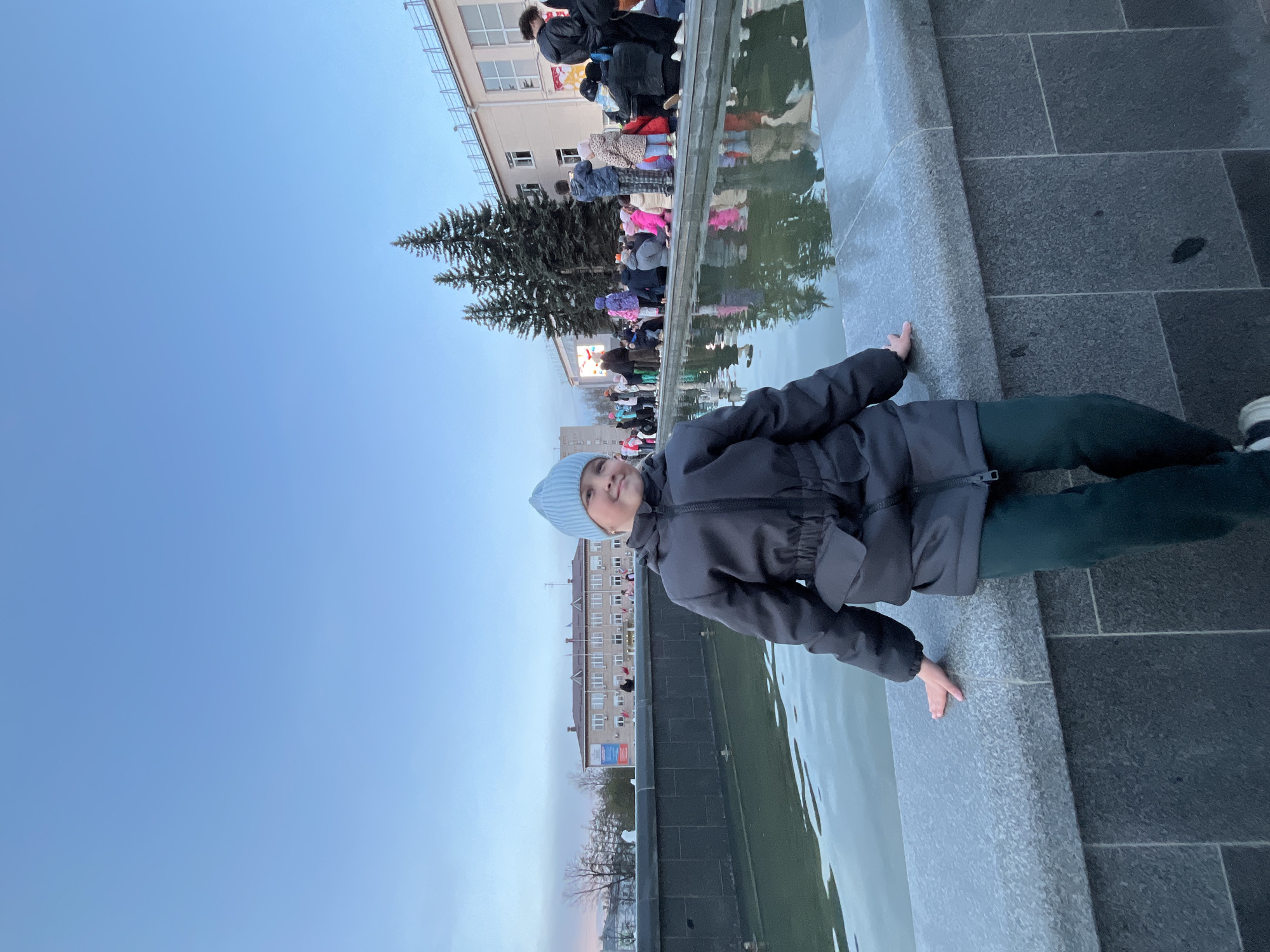 The image is a low-angle shot of a girl wearing a light blue hat and a gray jacket, lying prone with her hands touching the edge of a stone water feature or fountain. She is positioned along the horizontal axis, gazing upwards to her left. The body of water reflects the sky and nearby buildings and trees. In the background, there are multiple people standing near the water's edge or on a slightly elevated surface. The sky is a gradient of blue, transitioning from a lighter hue near the horizon to a deeper shade above. Buildings of several stories are visible in the background. There are coniferous trees to the left of the girl, and a bare tree near the lower left corner. The location is in Chaikovsky, Russia.