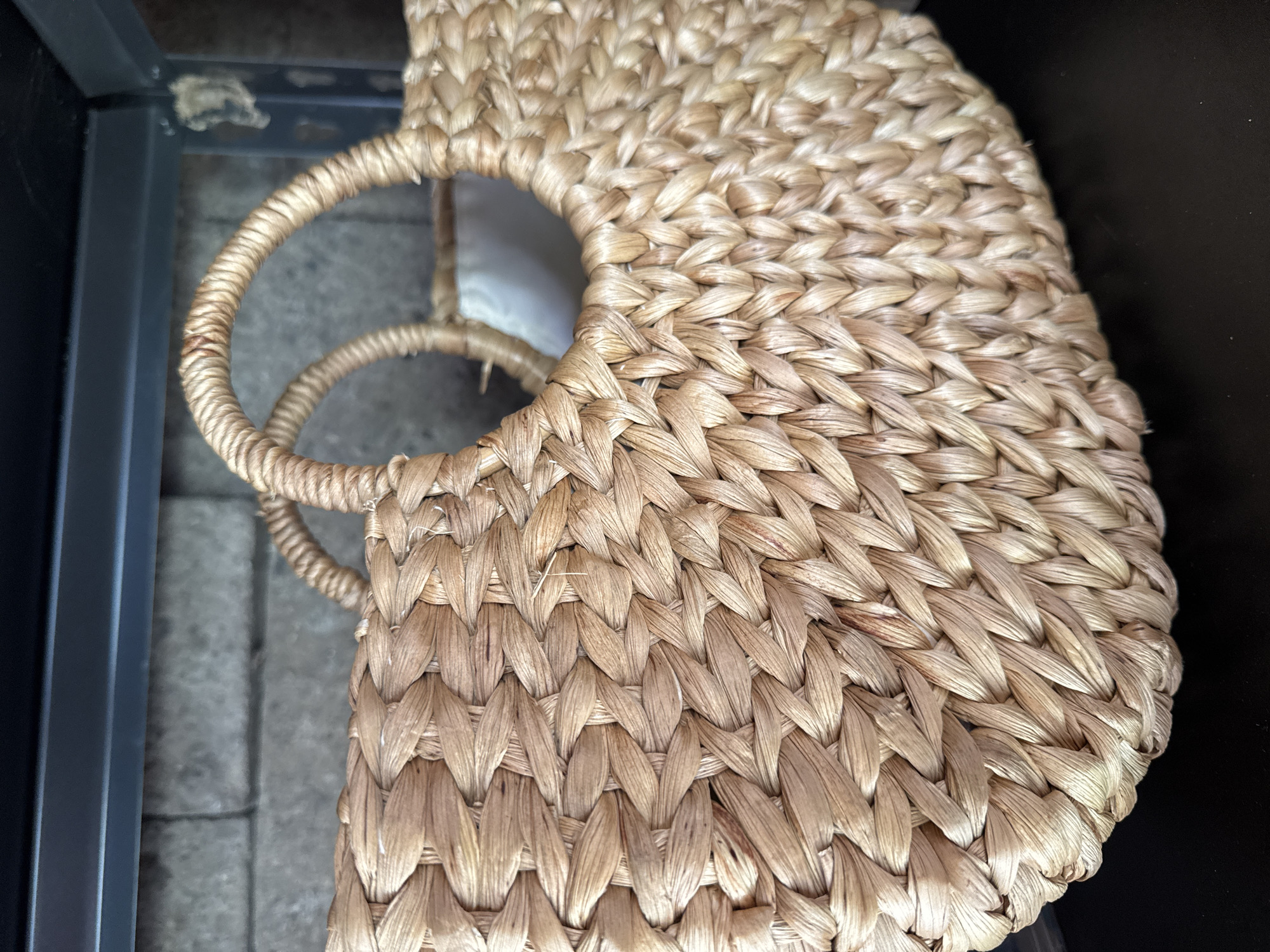 The image shows a tightly woven, light brown basket with two round handles. The basket appears to be resting indoors. The left edge of the frame includes a section of a dark-colored table or shelf with a tile floor below. The right edge shows a dark, possibly black, background. There are no people visible. The location, Kuta Selatan, Indonesia, cannot be determined from the visual information provided.