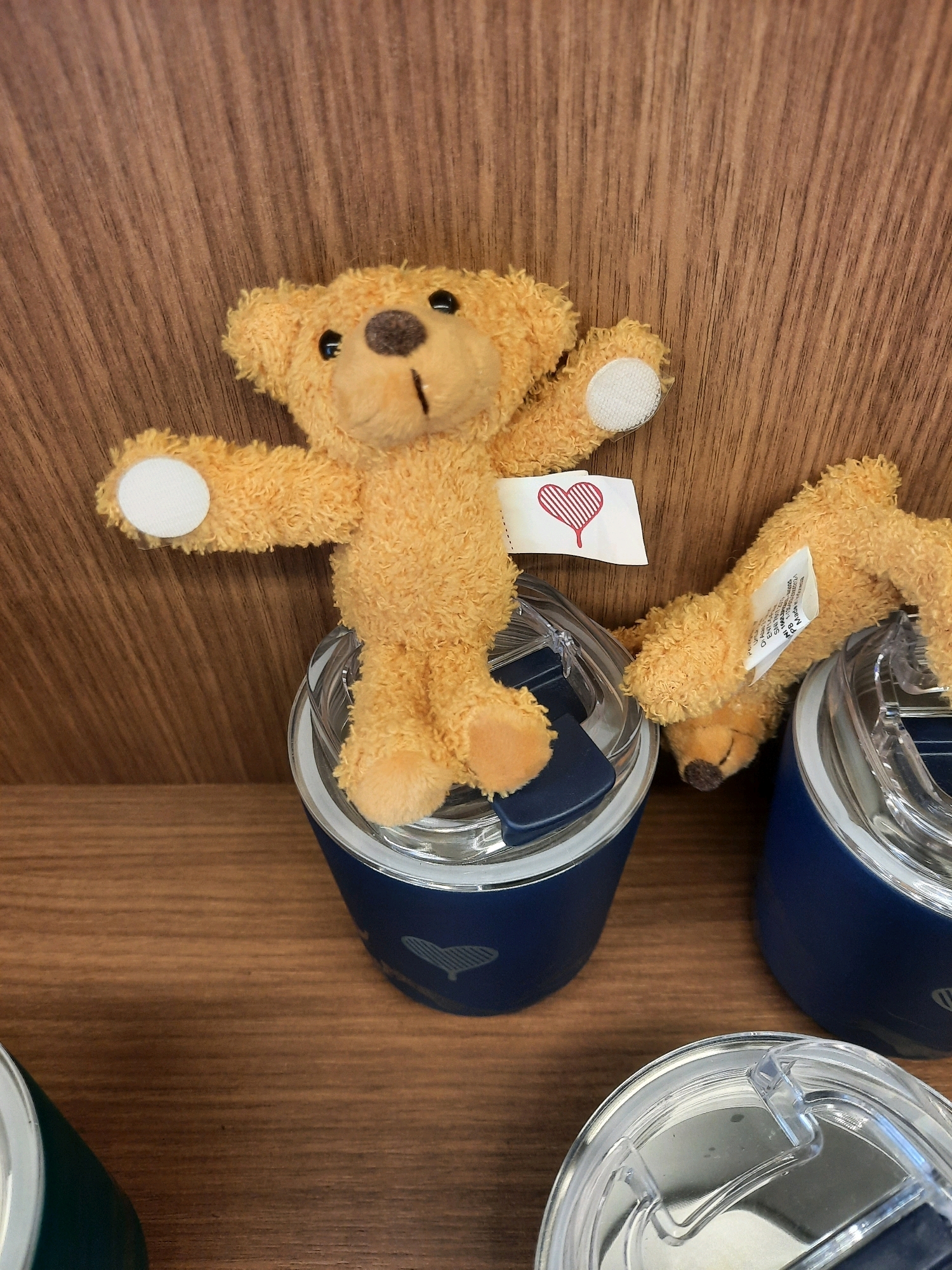 A small, fuzzy, orange teddy bear with black button eyes and a brown nose stands upright on top of a blue tumbler with a clear lid. The bear has its arms outstretched to the sides, and a white tag with a red heart design is attached to its right arm. Behind the bear and tumbler is a wooden surface with visible grain. Another blue tumbler with a clear lid is partially visible to the right of the first, and a second, similarly colored teddy bear is lying on its side on top of this tumbler. A third tumbler, dark green in color, is visible in the lower left corner of the image. The setting appears to be a retail environment, possibly a store display.