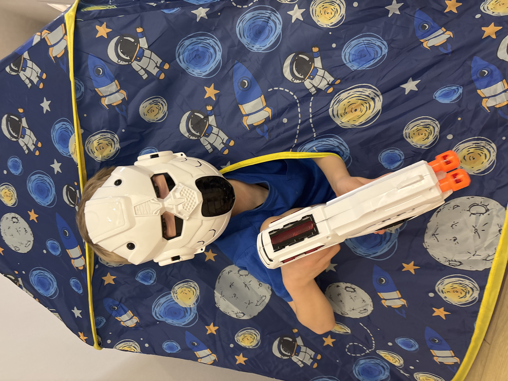A young child wearing a white robotic mask and a blue t-shirt is lying inside a space-themed play tent, holding a white and orange toy gun. The tent is patterned with images of astronauts, rockets, stars, and planets on a dark blue background. The tent's edges are trimmed with yellow. The image appears to have been taken from above. The location of the scene is unknown.