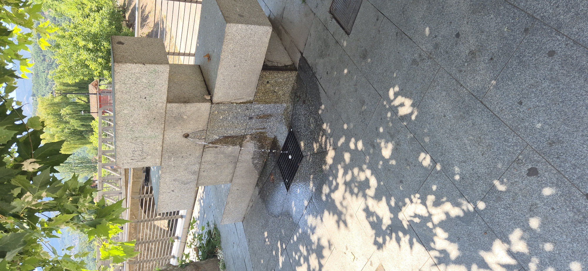 This media file depicts an outdoor scene, likely in Navaluenga, Spain, featuring a stone fountain structure. Water streams from a small spout onto the granite steps, creating a wet surface that reflects the dappled sunlight filtering through overhead foliage. The ground is paved with large, grey granite tiles, and a black grate is visible near the base of the fountain, likely serving as a drainage point.
In the background, lush green trees and a distant hillside suggest a natural, possibly mountainous, landscape. A small building with a red roof is partially visible amongst the trees, hinting at a park or public area. The sunlight filtering through the leaves casts distinct shadows on the paved ground, indicating it is daytime, likely sunny. There are no people visible in the frame, focusing the attention on the architectural elements and the natural surroundings.
The stone structure appears to be a multi-tiered fountain or a decorative water feature integrated into a walkway or plaza. The overall impression is one of tranquility and a connection with nature, enhanced by the presence of water and greenery. The lighting and the shadows suggest a bright, clear day. No visible text is present in the image.