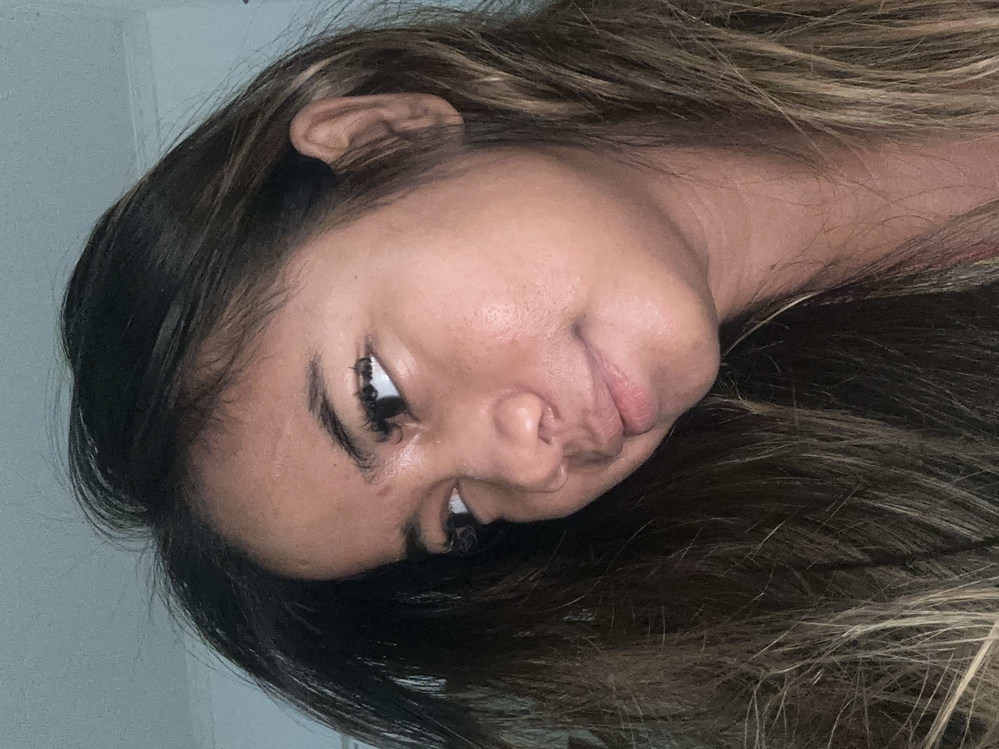 The image shows a close-up, shoulder-up view of a young woman with long, dark brown and blonde streaked hair. She is looking to her left, with her head tilted slightly down. Her skin appears to have a light sheen, and she is wearing subtle makeup, including mascara. The background is a plain, light blue wall with a white molding at the top. The lighting suggests an indoor environment, possibly a home or studio. There is no information in the image that indicates the location in Sukawati, Indonesia.
