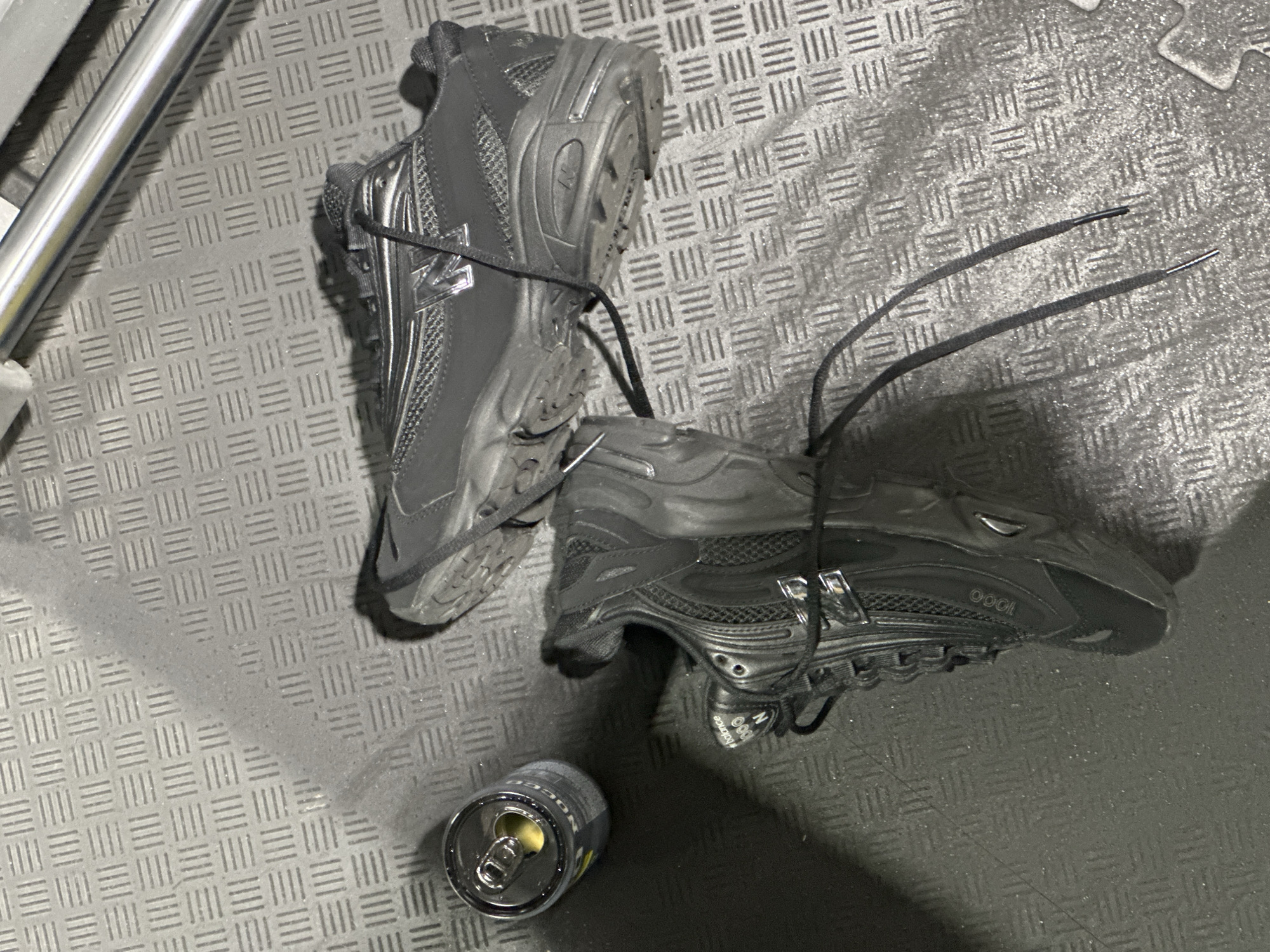 The scene captures an overhead view of an indoor setting with a dark grey, textured floor, likely rubber matting, featuring a grid pattern of raised parallel lines. Two black New Balance athletic shoes are present. One shoe, identified as a New Balance 1000 model, rests upright on the left with its laces untied. The other shoe is inverted, positioned on its sole and side, towards the upper right, also with untied laces. Both shoes display a metallic silver 'N' logo. To the upper left of the upright shoe, an opened, dark-colored beverage can with a silver pop-top is visible, containing a yellowish liquid. A horizontal silver metallic bar or pipe is partially visible in the upper right corner of the frame. No individuals are depicted. The specific location within Tunbridge Wells, United Kingdom, cannot be determined from the visual evidence, though the environment suggests a gym or leisure facility.