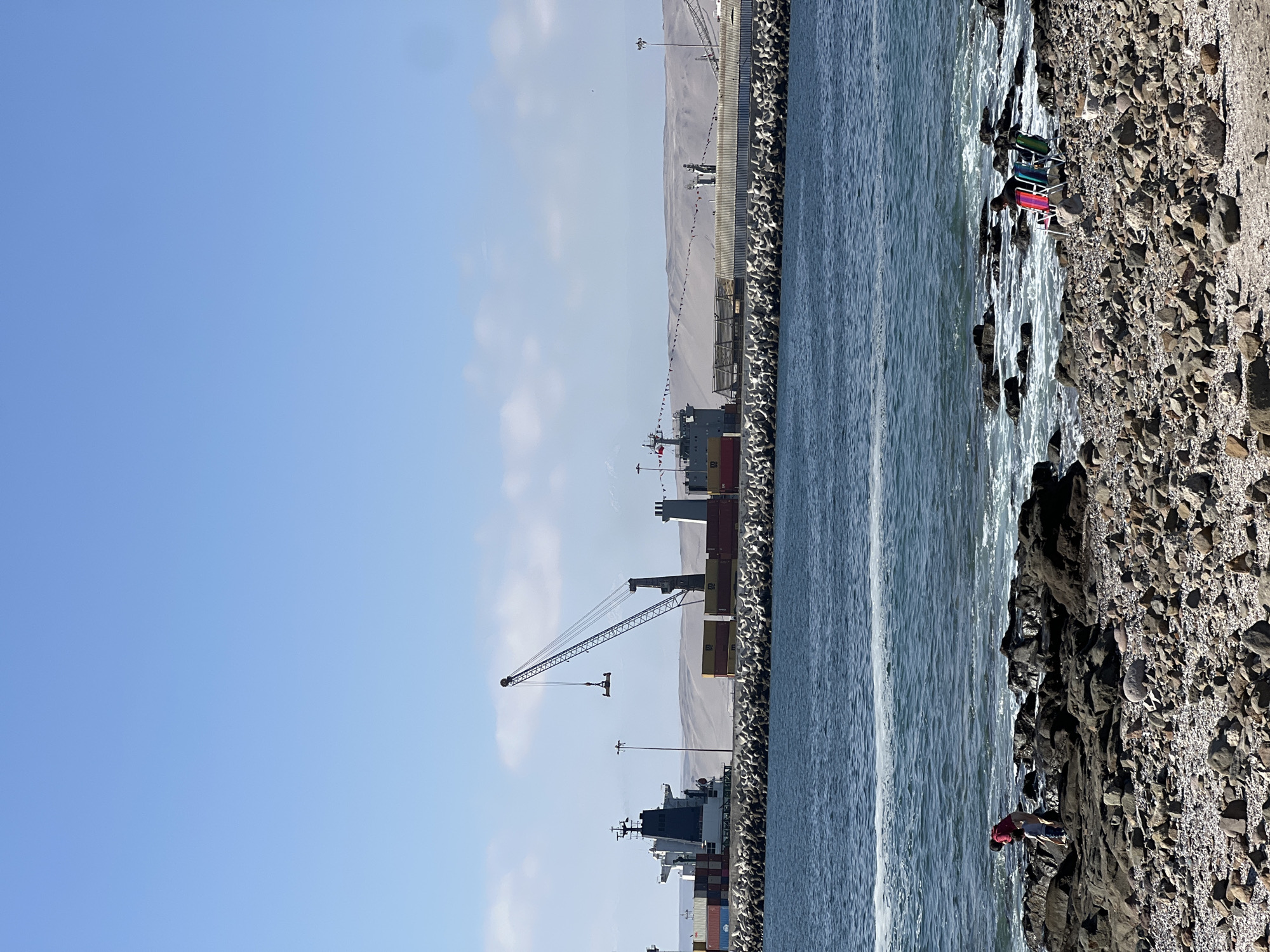 The image shows a view of the Port of Arica in Chile. Several cargo ships are docked along a breakwater. A large crane is present, presumably for loading and unloading containers. The landscape behind the port appears to be a desert. The foreground consists of a rocky beach along the shoreline of the ocean. There are some chairs set up on the beach. Also in that area, a person is bending over or squatting near the rocks.